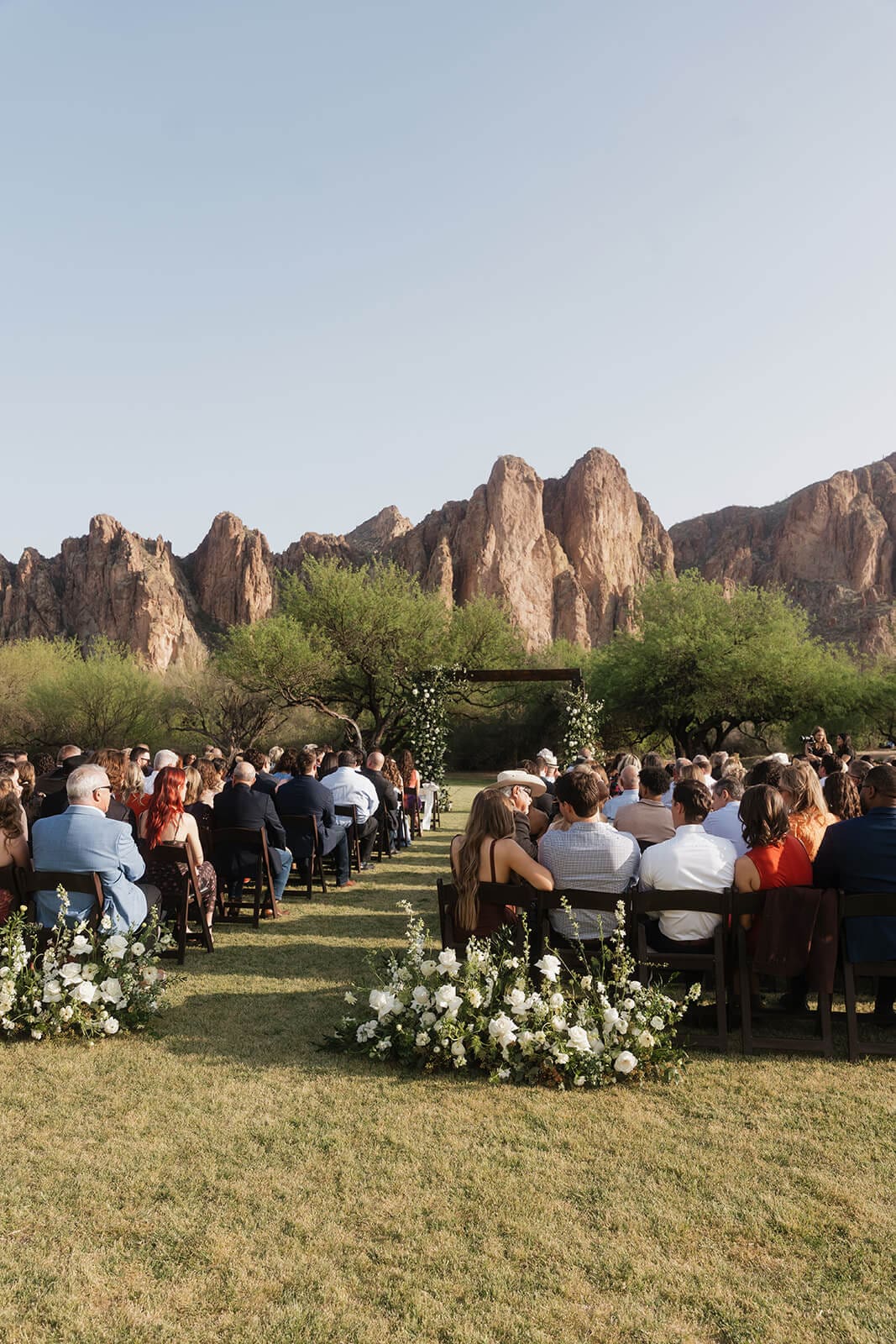 Outdoor wedding ceremony in progress on the lawn with guests seated and a floral arch at the altar against the rocky cliffs at Saguaro Lake Guest Ranch wedding venue in Mesa Arizona
