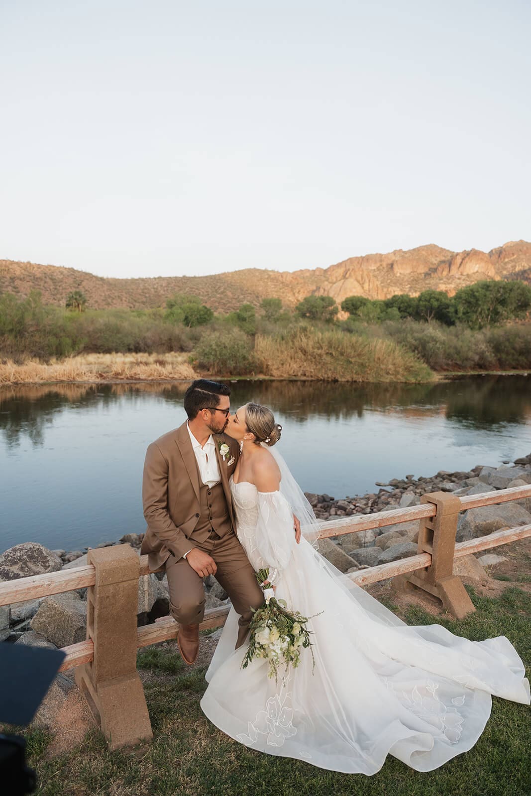 Bride and groom sitting together on a wooden bridge kissing with the Salt River and rocky cliffs in the background at Saguaro Lake Guest Ranch wedding venue in Mesa Arizona
