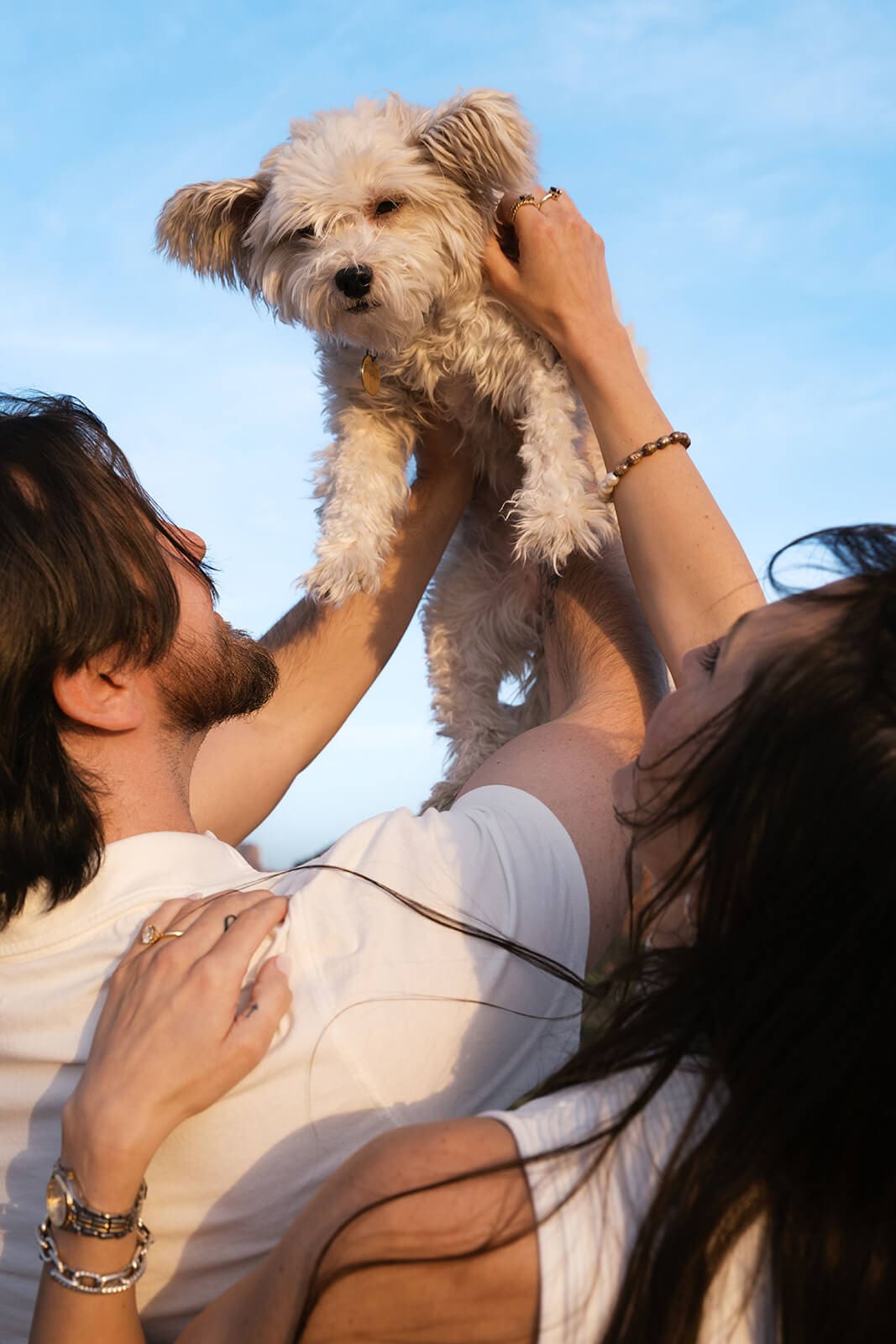 A woman holds a small fluffy white dog up in the air against a blue sky, a man's hand resting on her back, both dressed in white.
