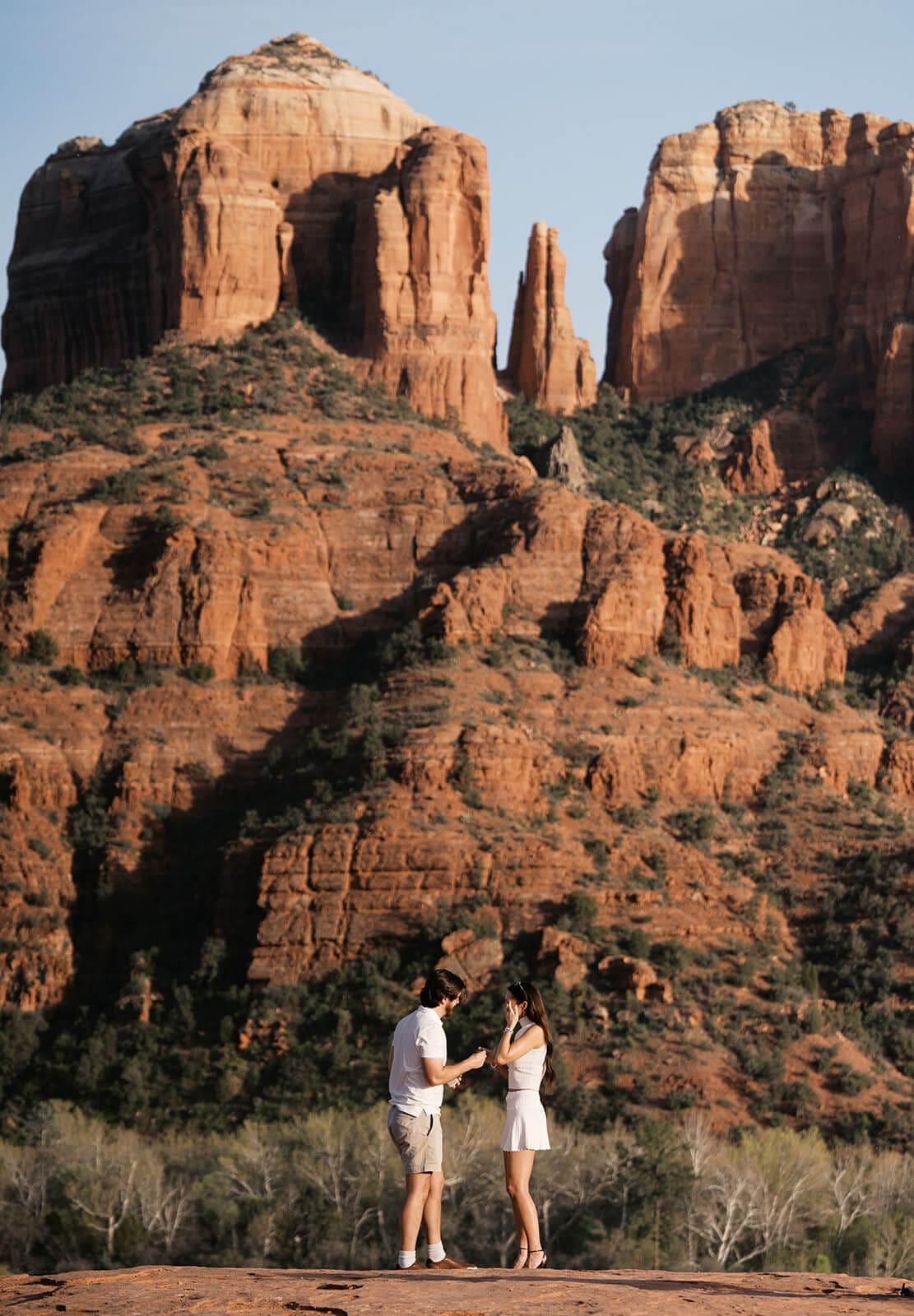 A couple stands on a red rock overlook in Sedona, Arizona, the man sliding a ring onto the woman's finger with Cathedral Rock's towering sandstone formations glowing in the background.
