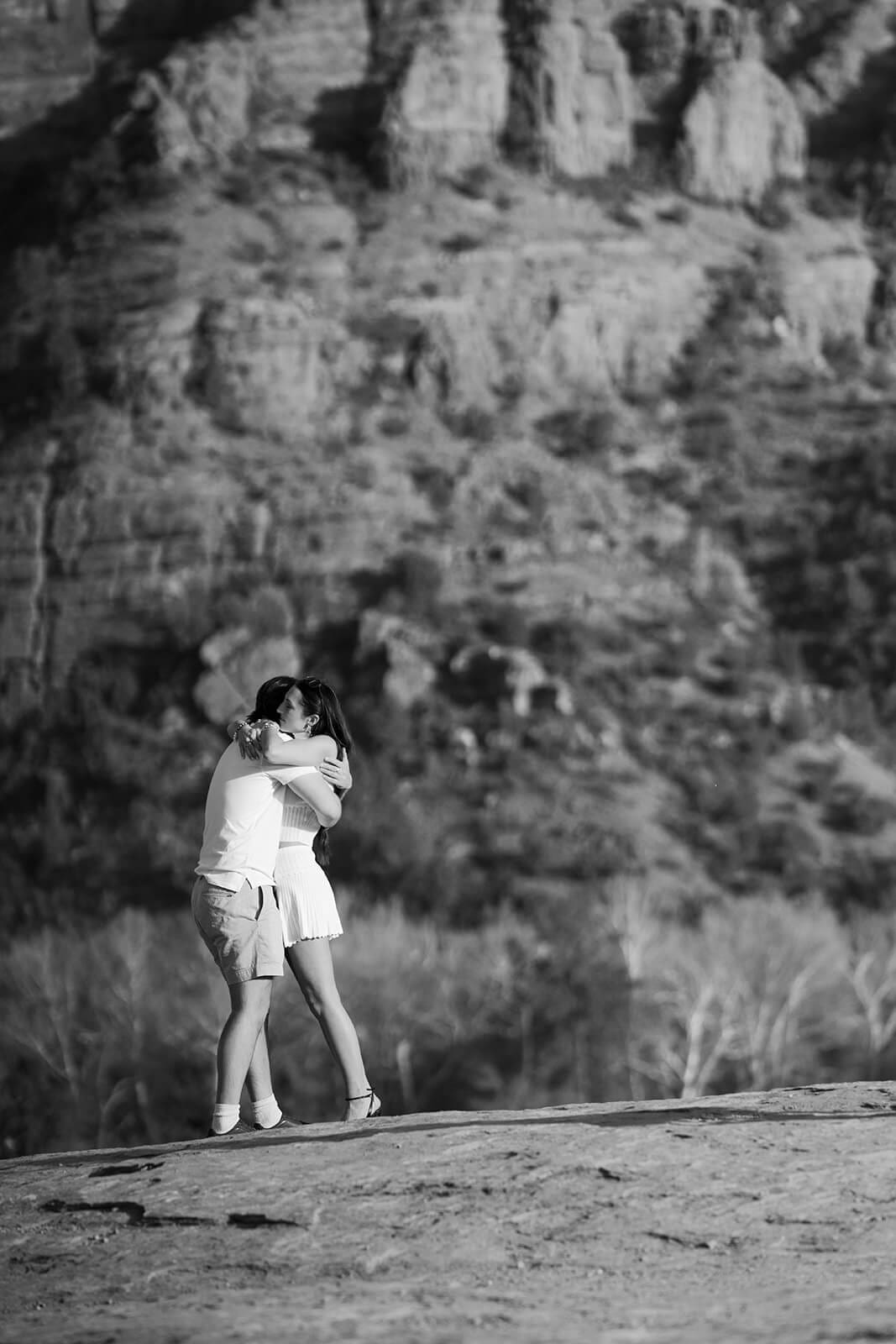 Black and white photo of a newly engaged couple embracing on a flat rock outcropping in Sedona, with dramatic textured cliff faces filling the background.

