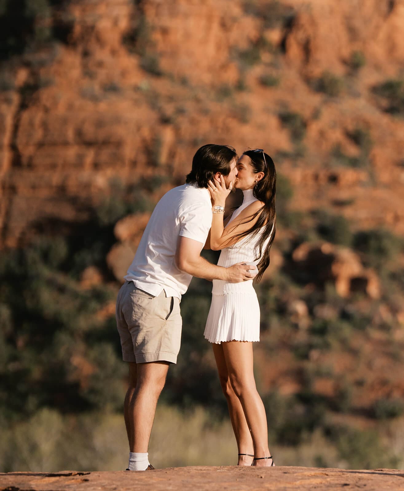 A couple shares a kiss on a red rock overlook in Sedona, both dressed in white, with warm sandstone cliffs behind them.
