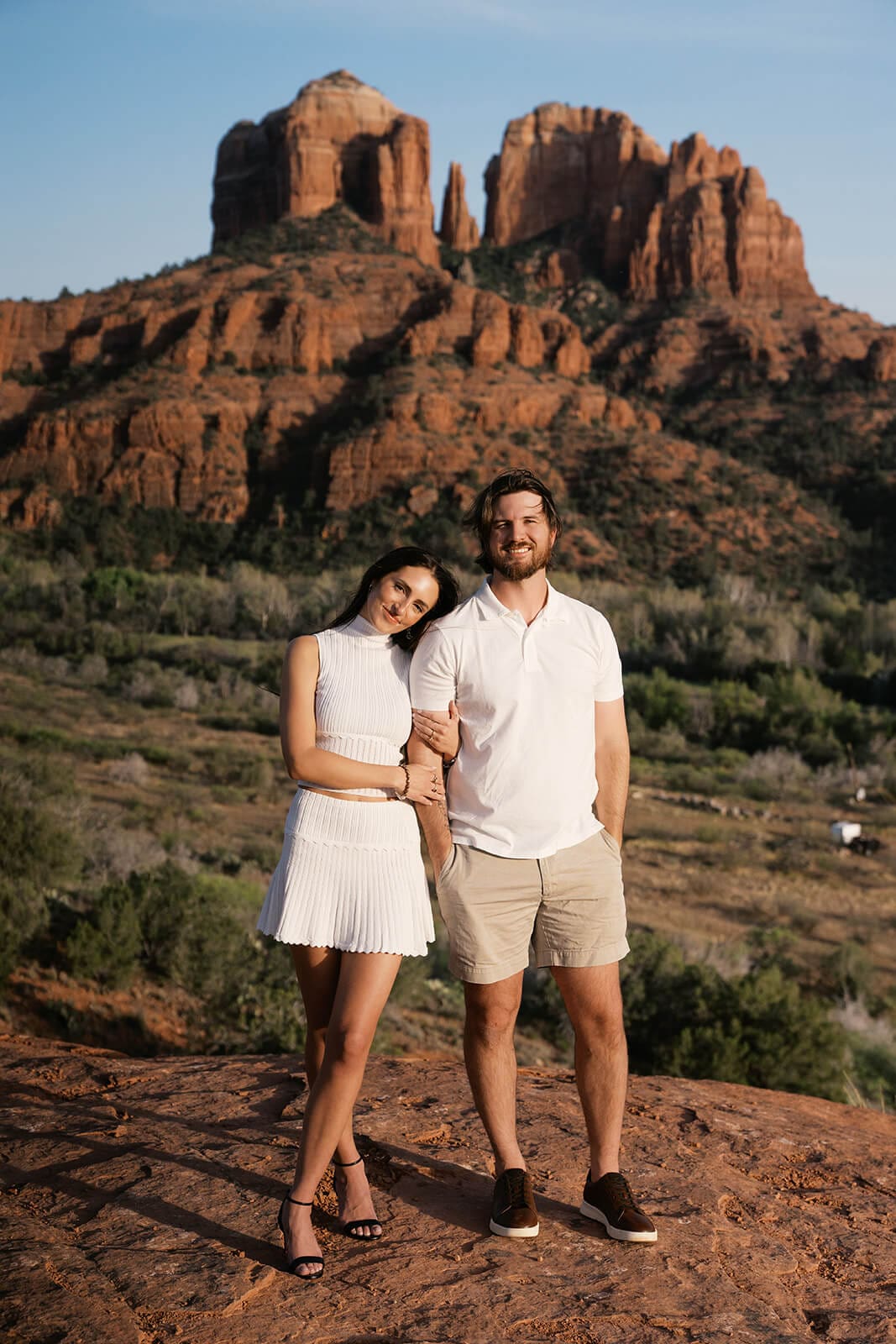 A newly engaged couple poses together on a red rock formation in Sedona, both dressed in white and cream tones, with Cathedral Rock rising behind them in the golden hour light.
