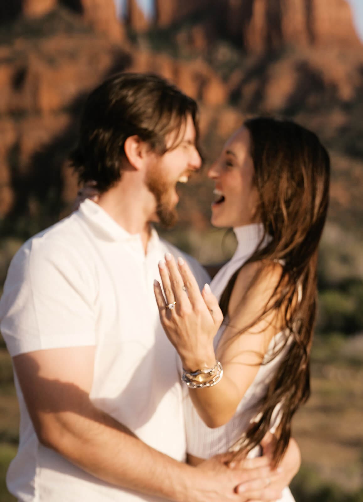 A newly engaged couple laughs joyfully together, the woman's hand with a diamond ring pressed against the man's chest, with warm Sedona red rock formations softly blurred in the background.