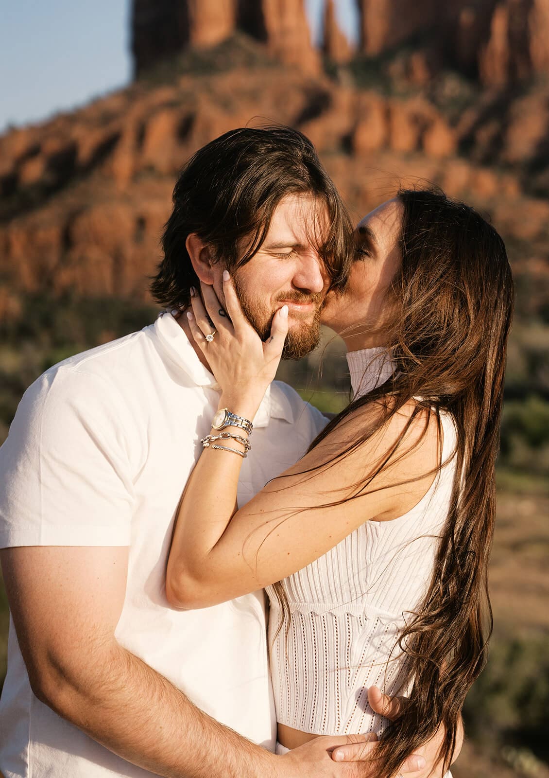 Close-up of a couple sharing a kiss outdoors, the woman's hand gently cradling the man's face, with Sedona's red rock formations softly blurred behind them.
