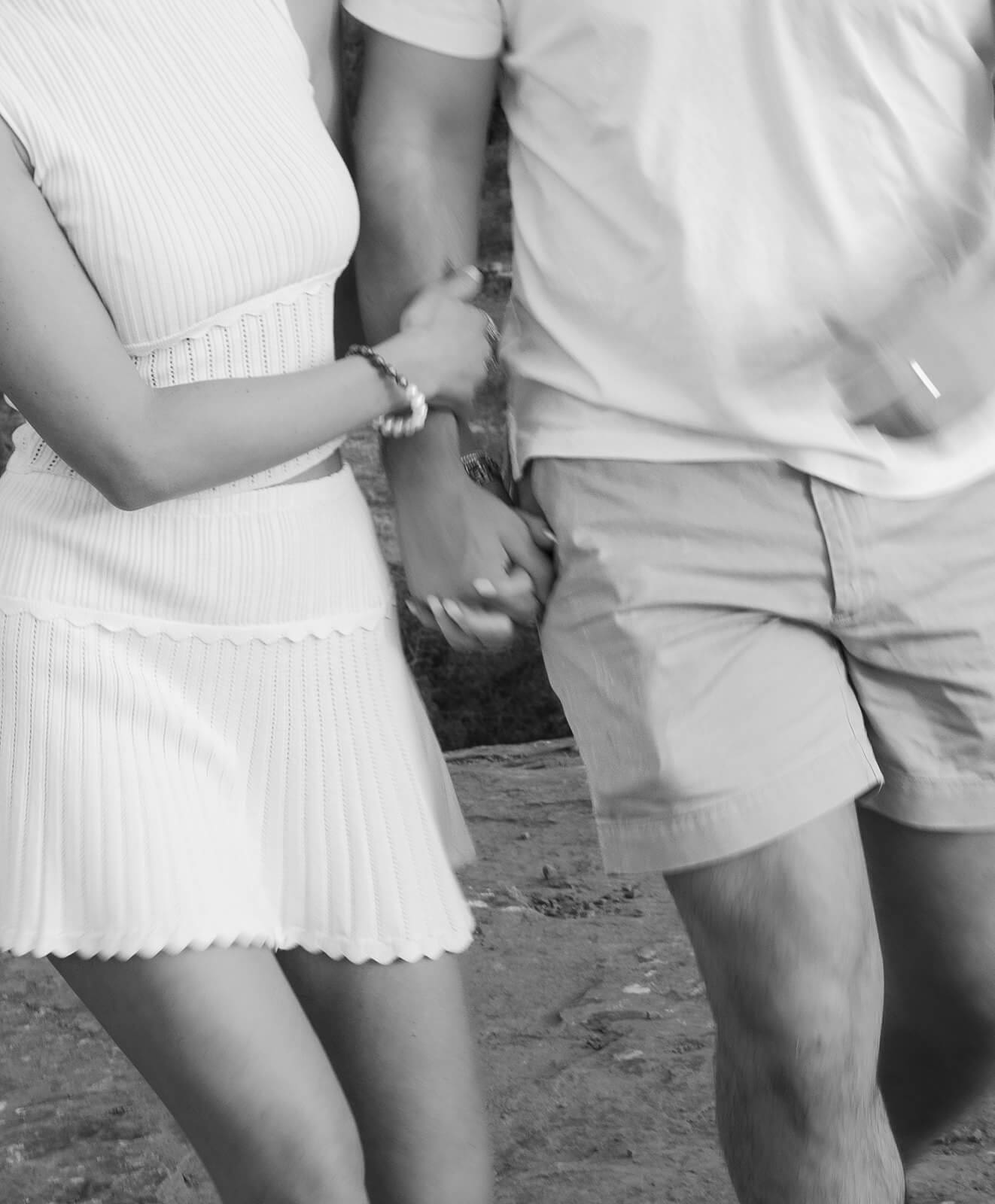 Black and white close-up of a couple holding hands, framed from the waist down, both dressed in white and standing on a rocky surface.
