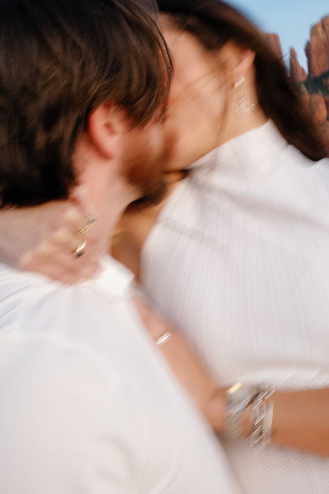 Close-up of a couple sharing a kiss, the woman's hand with her engagement ring and bracelet resting on the man's chest, with warm Sedona red rocks softly blurred in the background.
