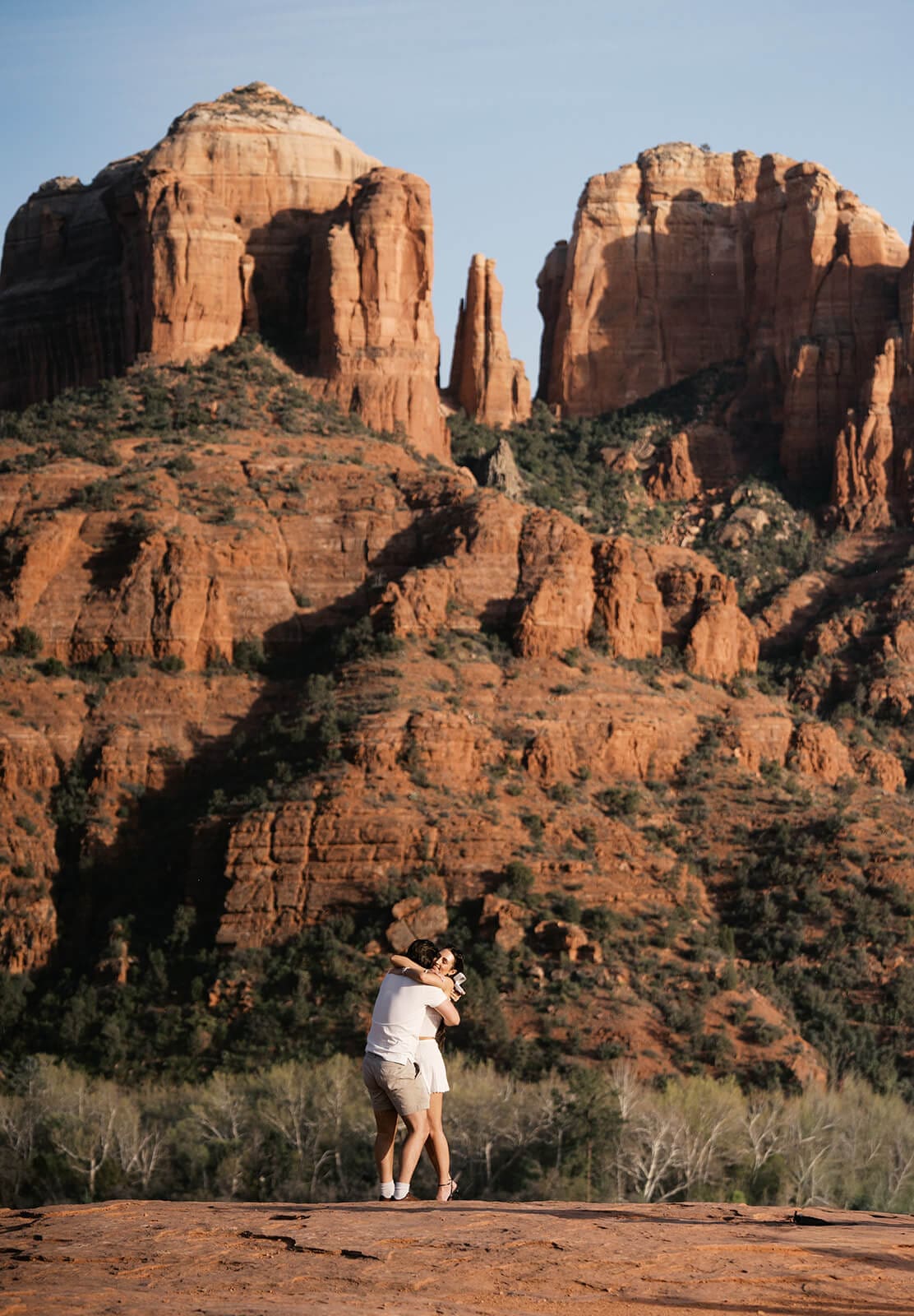 A wide shot of a woman dancing alone on a red rock outcropping in Sedona, with Cathedral Rock's towering formations filling the background.
