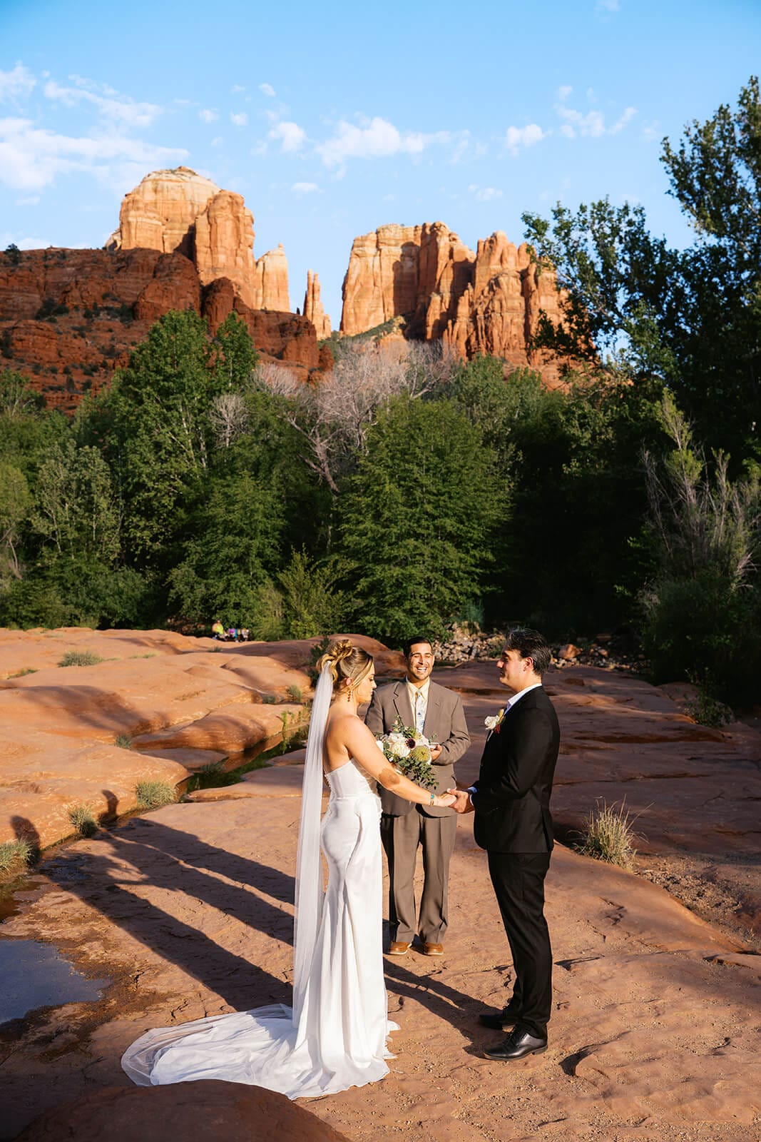 Bride and groom standing together on red rocks during their Sedona elopement, with Cathedral Rock and desert trees behind them.
