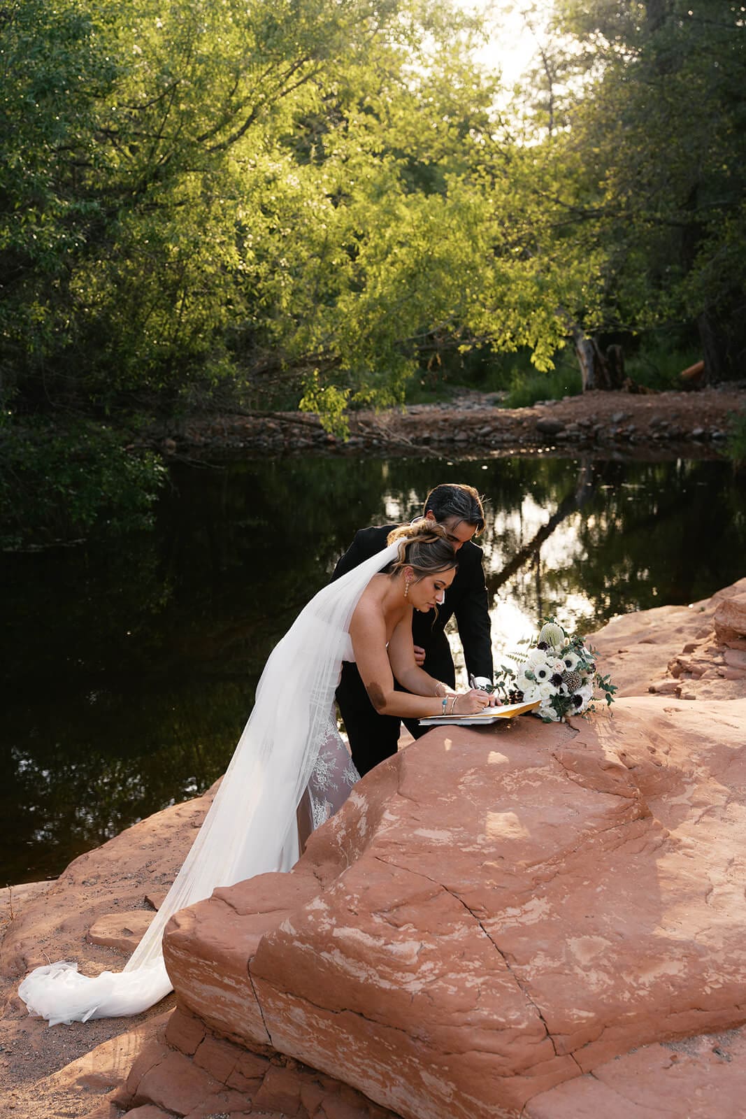 Bride and groom signing their marriage license after their Sedona elopement ceremony beside the creek, with vow books and flowers nearby.