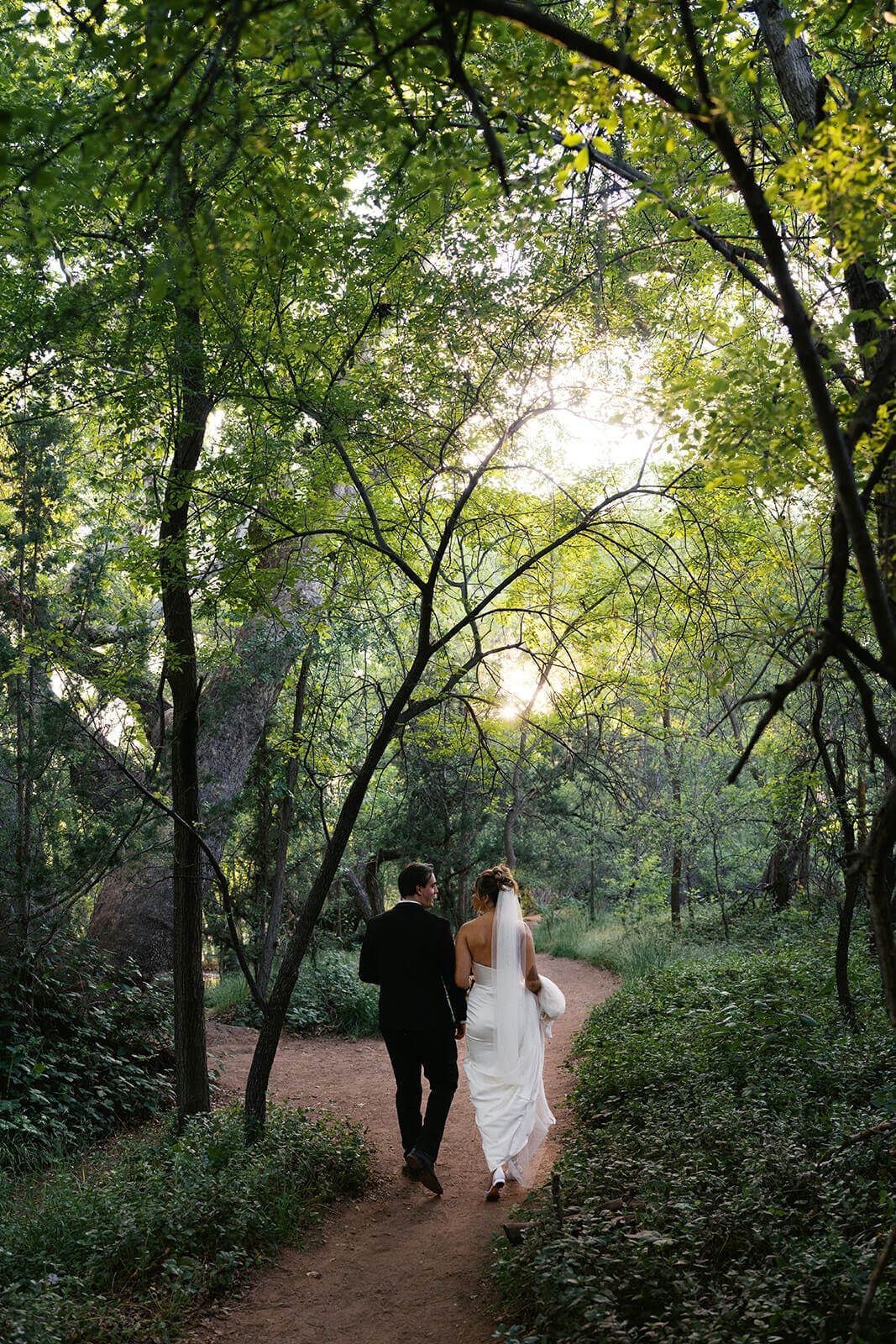 Newlyweds walking hand in hand through a wooded trail after their Sedona elopement ceremony, surrounded by trees and soft light.