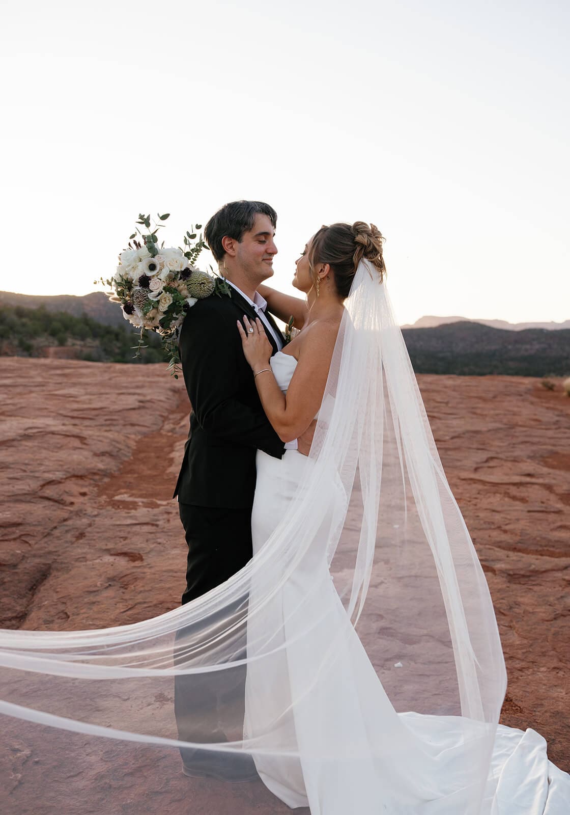 Bride and groom embracing on the red rocks during their Sedona elopement, with her long veil trailing behind them in the desert breeze.