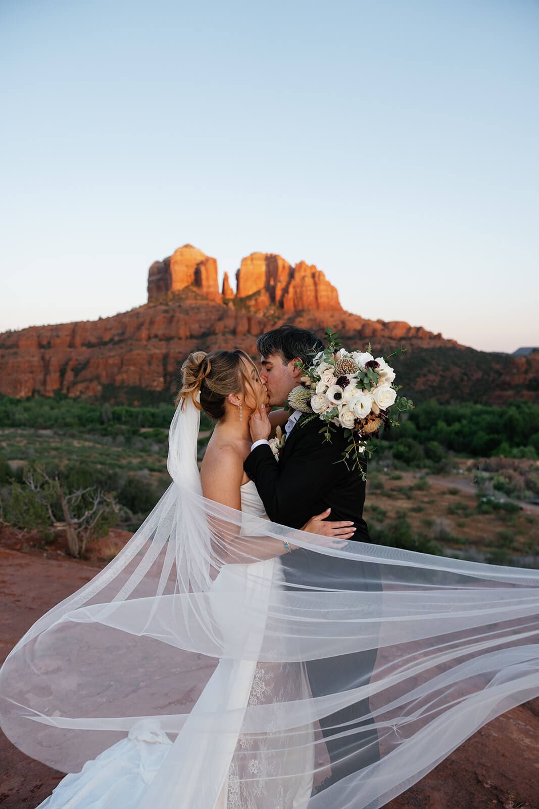 Bride and groom kissing during their Sedona elopement portraits, with her long veil blowing across the frame in front of Cathedral Rock.