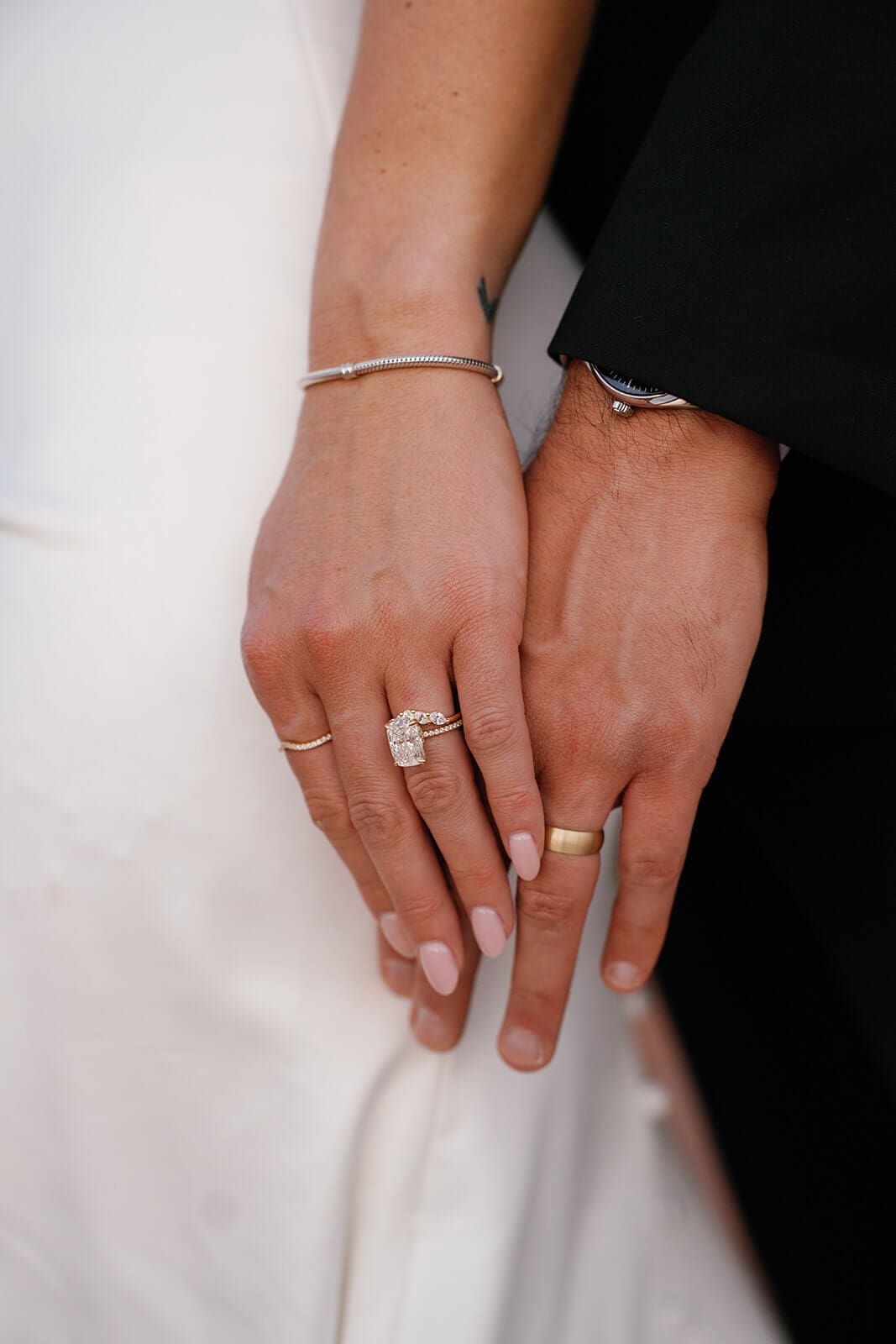 Close-up of the bride and groom’s hands showing their wedding rings after their elopement, with her white dress and his black suit in the background.