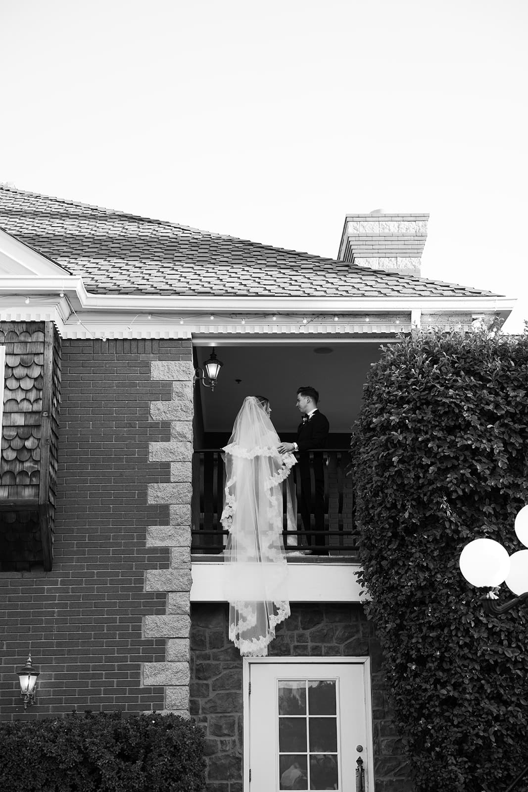 Bride standing on a second-story balcony of the ivy-covered manor house at Stonebridge Manor, photographed in black and white.