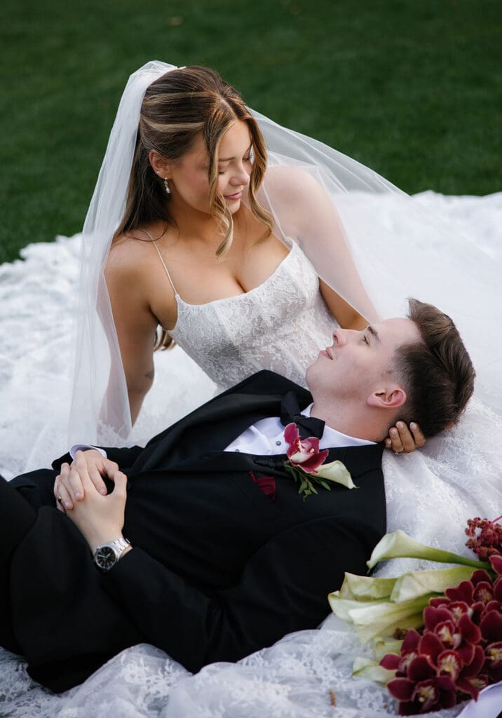 Bride leaning over the groom as he lies on the grass beside her wedding dress and colorful bouquet.