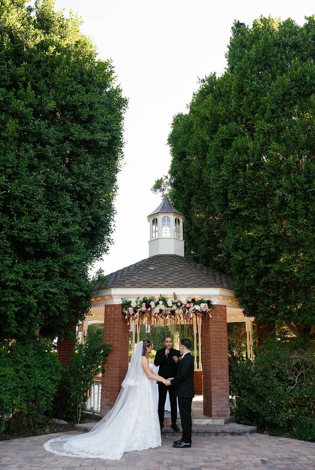 Bride and groom exchanging vows in front of the brick gazebo surrounded by tall hedges and guests at Stonebridge Manor wedding venue in Mesa Arizona
