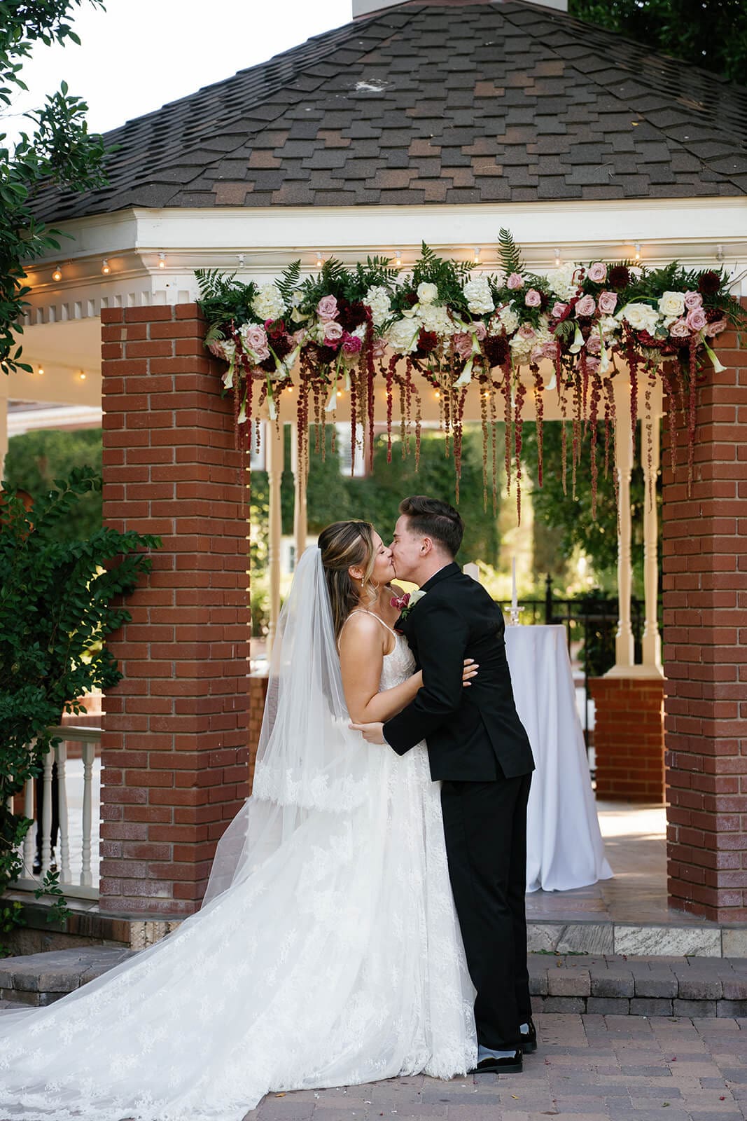 Bride and groom sharing their first kiss under a floral arch in front of the brick gazebo at Stonebridge Manor, an elegant garden wedding venue in Mesa Arizona
