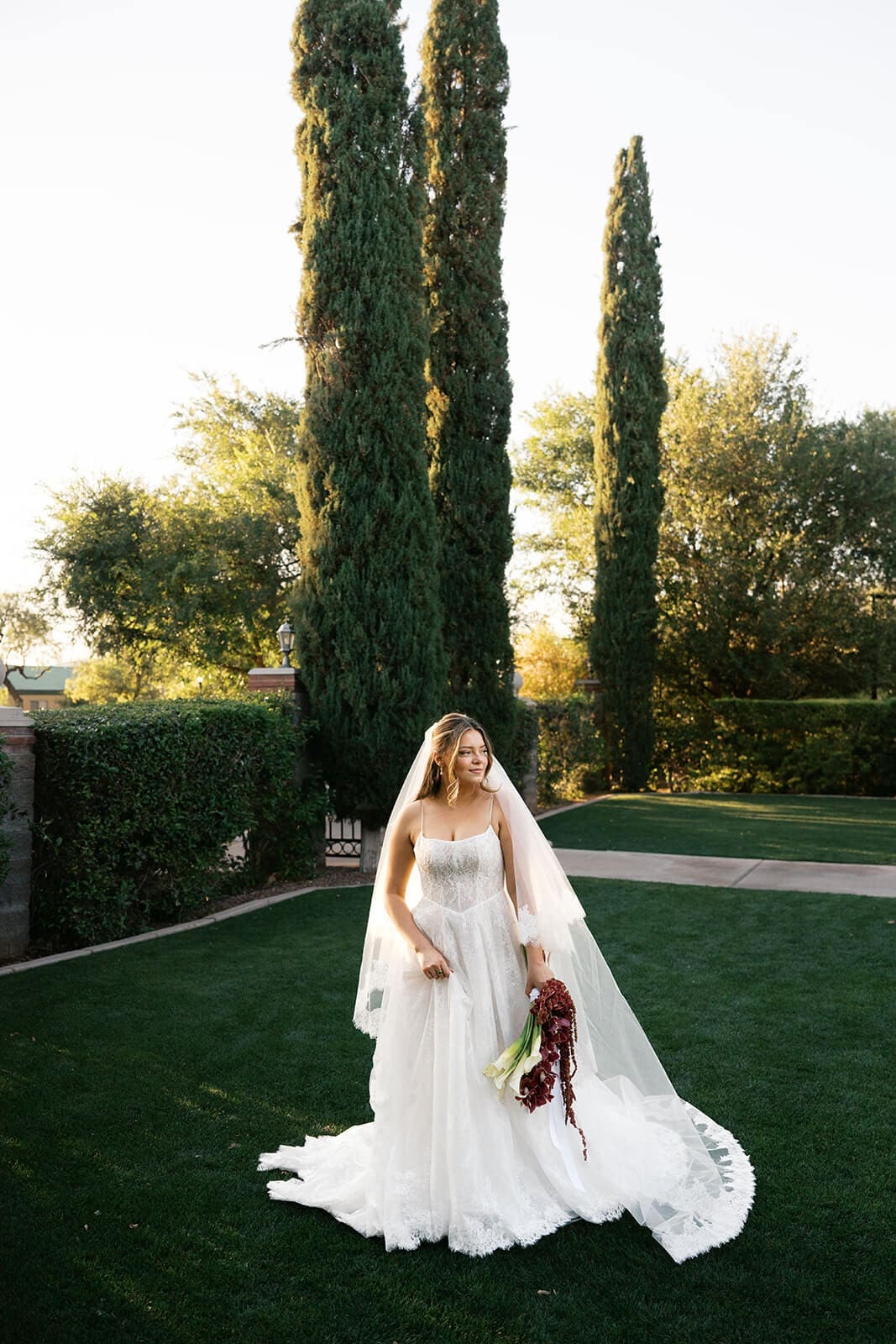Bride standing alone on the manicured lawn holding her bouquet with tall cypress trees behind her at Stonebridge Manor garden wedding venue in Mesa Arizona
