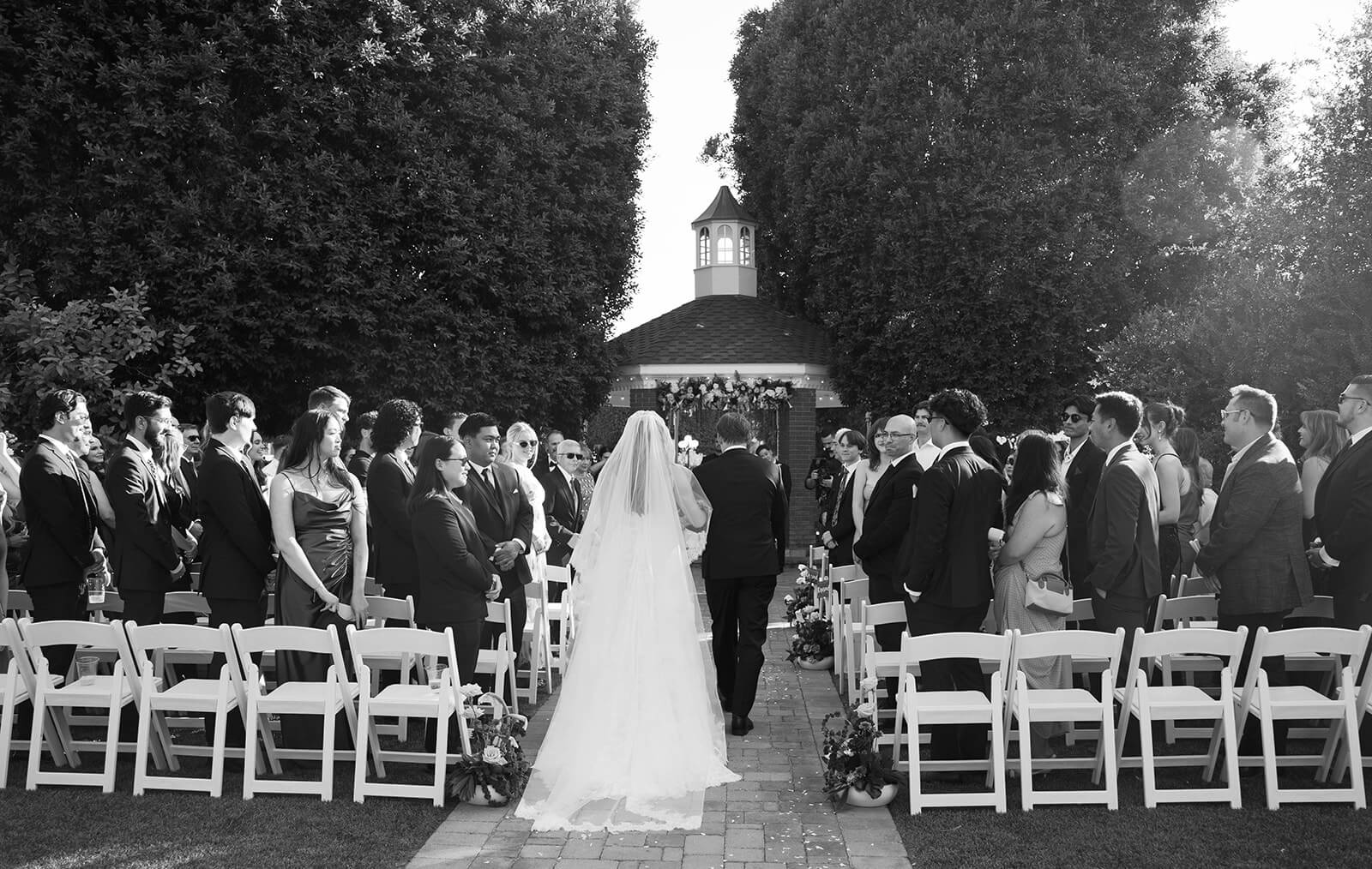 Black and white photo of bride walking down the aisle toward the gazebo with guests standing on either side at Saguaro Lake Guest Ranch wedding venue in Mesa Arizona
