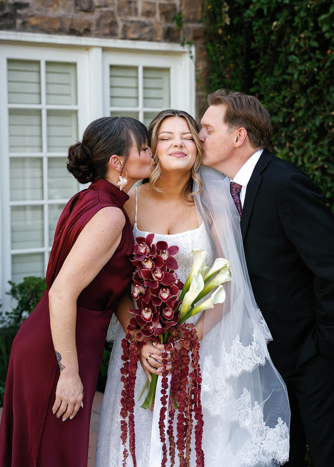 Bride standing between her parents while holding a cascading burgundy orchid bouquet during wedding portraits at Stonebridge Manor.