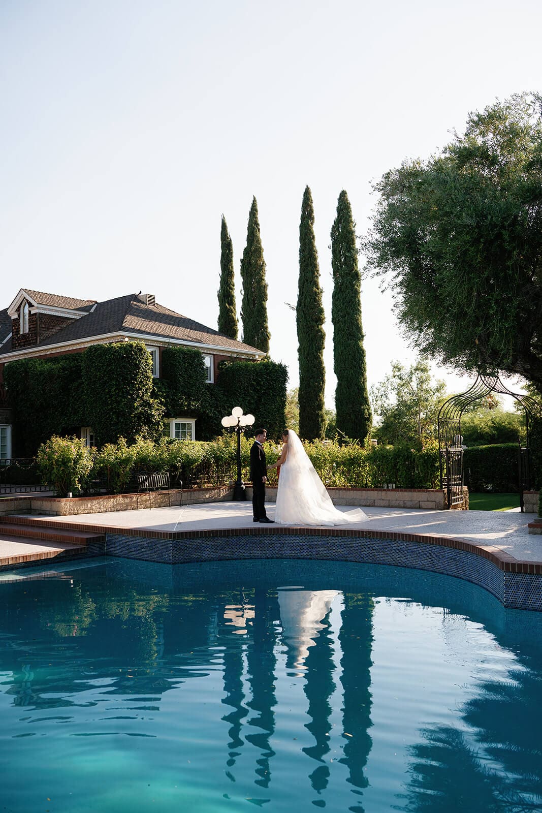 Bride and groom standing beside the blue-tiled pool at Stonebridge Manor with reflections of the venue and cypress trees visible in the water.