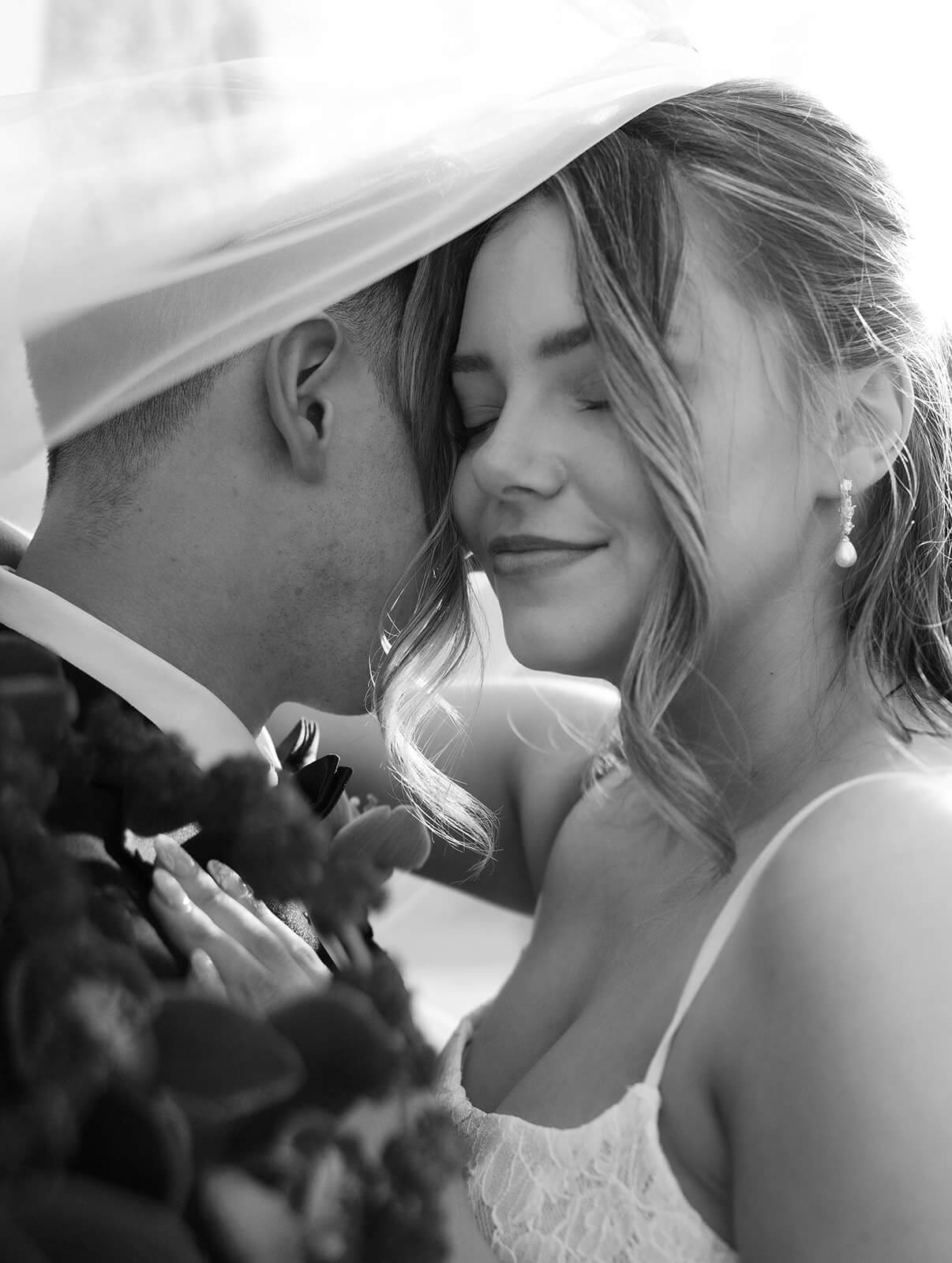 Black and white close-up portrait of the bride and groom embracing beneath the wedding veil during golden hour.