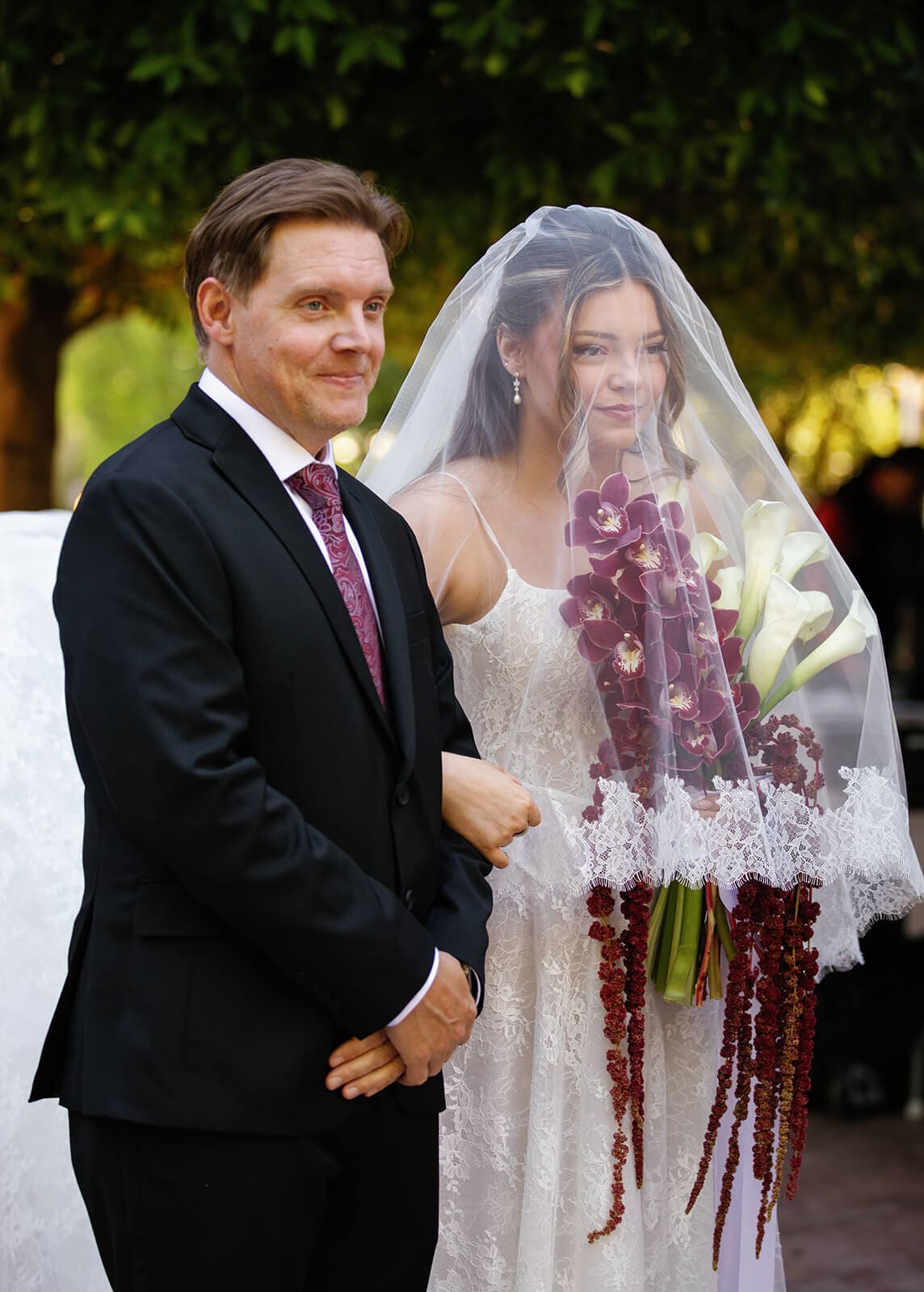 Bride holding a cascading burgundy orchid bouquet while standing beside her groom before their outdoor ceremony at Stonebridge Manor.