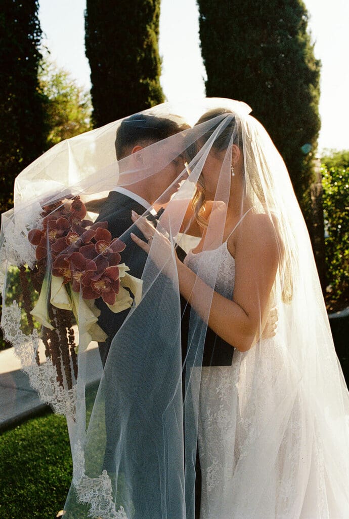Newlyweds kissing beneath the bride’s veil in warm backlit sunlight.