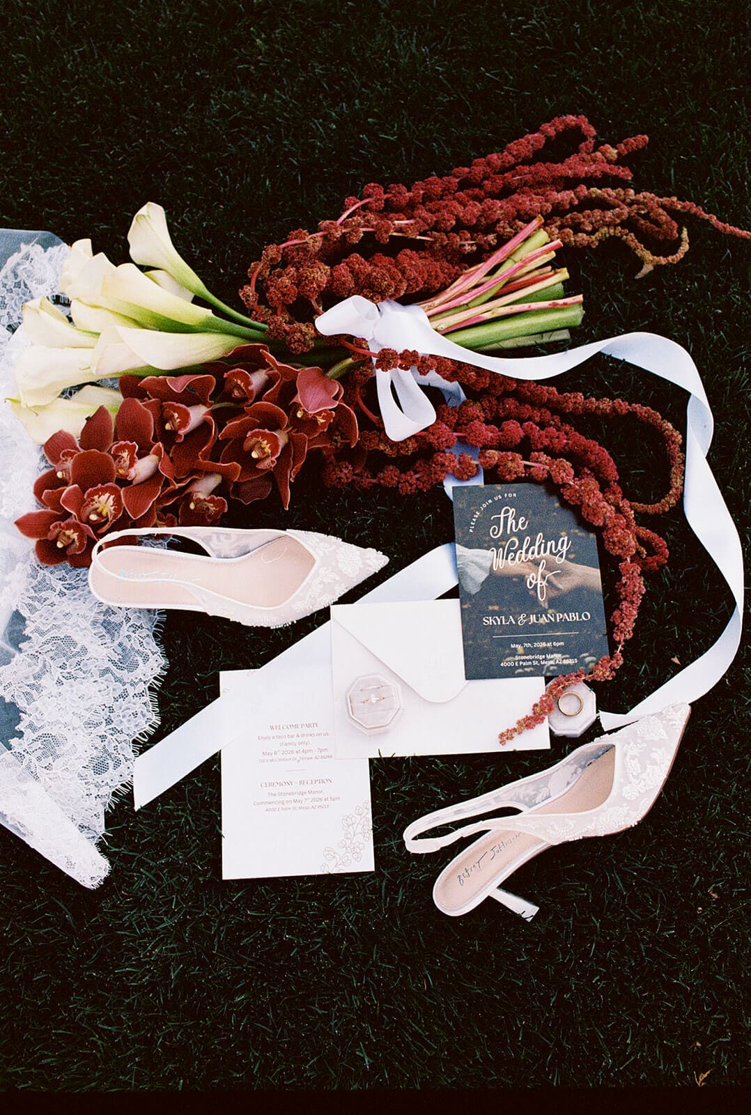 Flat lay of wedding details, including invitations, lace heels, wedding rings, and a burgundy orchid bouquet displayed on the lawn at Stonebridge Manor.