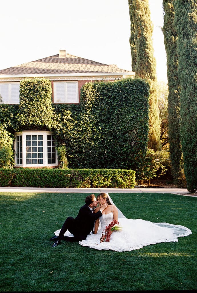 Bride and groom sitting together on a manicured lawn in front of an ivy-covered estate.
