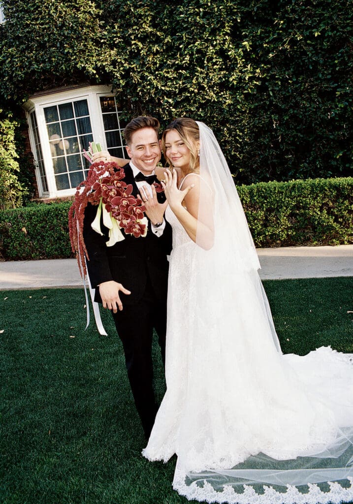 Bride and groom posing on a lawn in front of an ivy-covered estate, with the bride holding a burgundy bouquet.