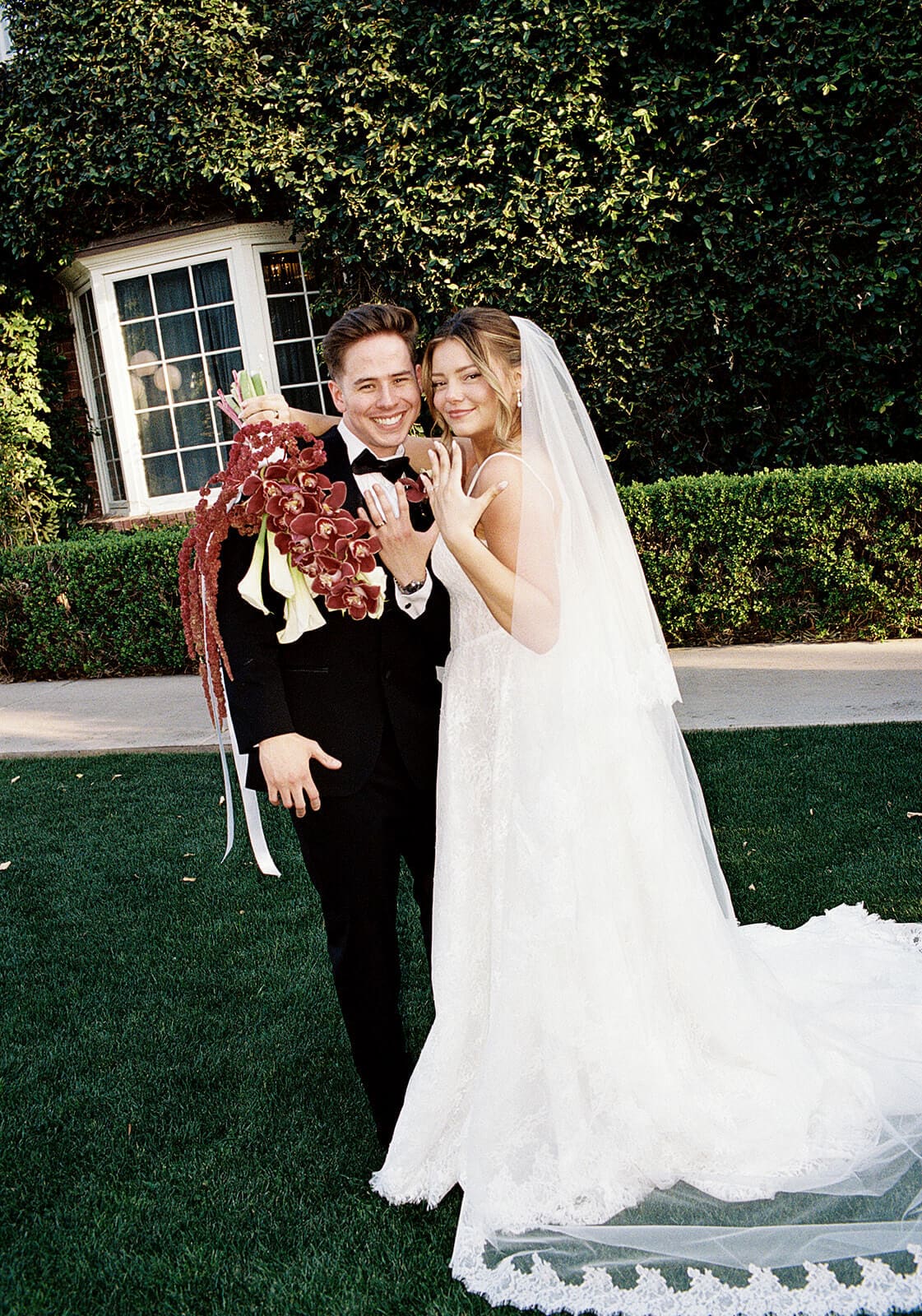 Bride and groom posing together in front of the ivy-covered manor house at Stonebridge Manor, holding a dramatic burgundy orchid wedding bouquet.