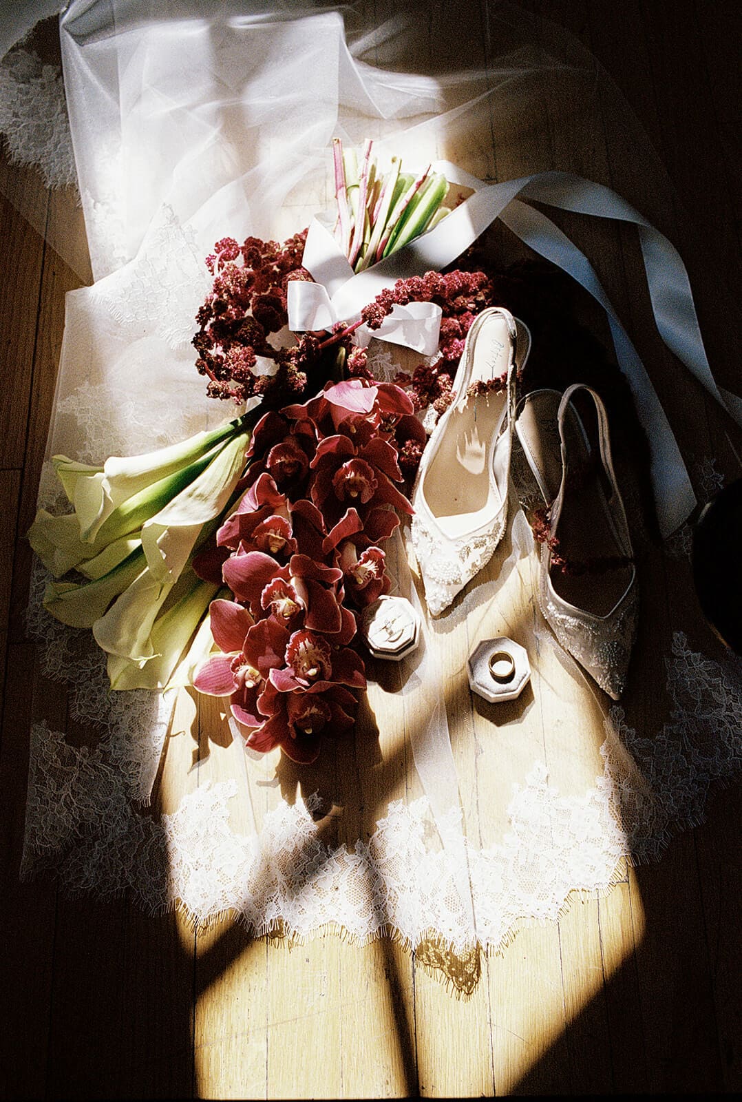 Wedding detail flat lay featuring a cathedral veil, burgundy orchids, calla lilies, wedding rings, and embroidered bridal shoes in natural window light.