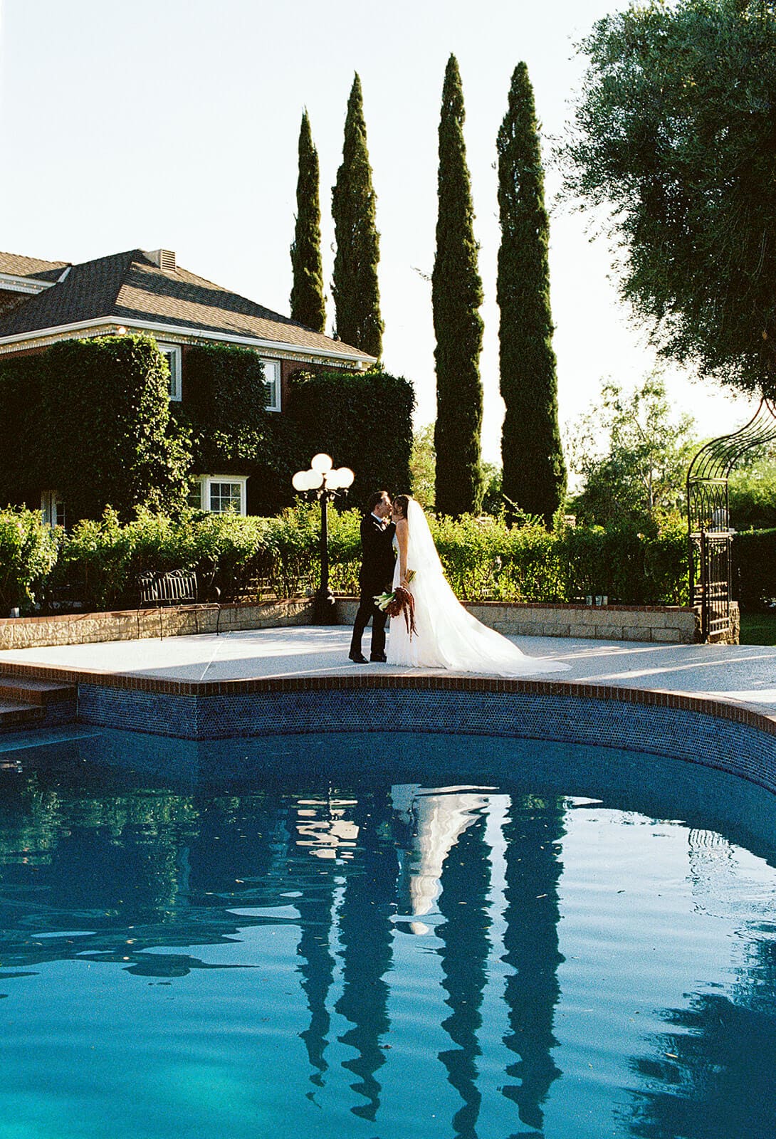 Bride and groom standing together reflected in the blue tiled pool with tall cypress trees behind them at Stonebridge Manor wedding venue in Mesa Arizona
