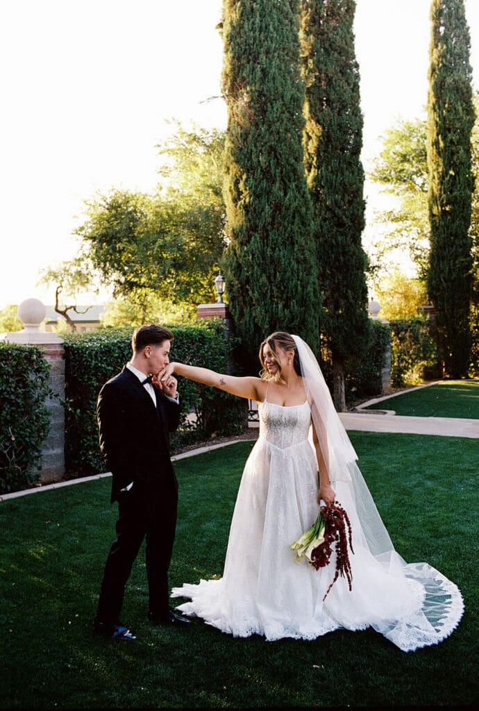 Bride reaching toward the groom while they stand together on a green lawn surrounded by tall cypress trees.