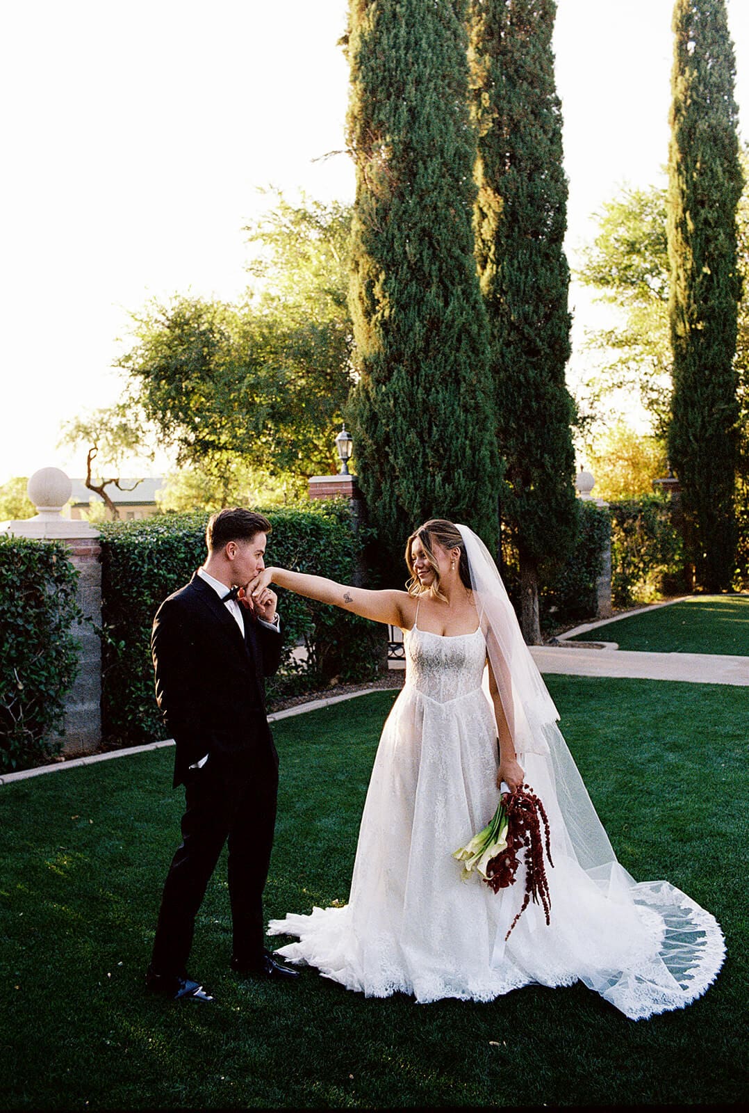 Bride and groom laughing together on a manicured lawn framed by tall cypress trees at Stonebridge Manor, an elegant garden wedding venue in Mesa Arizona