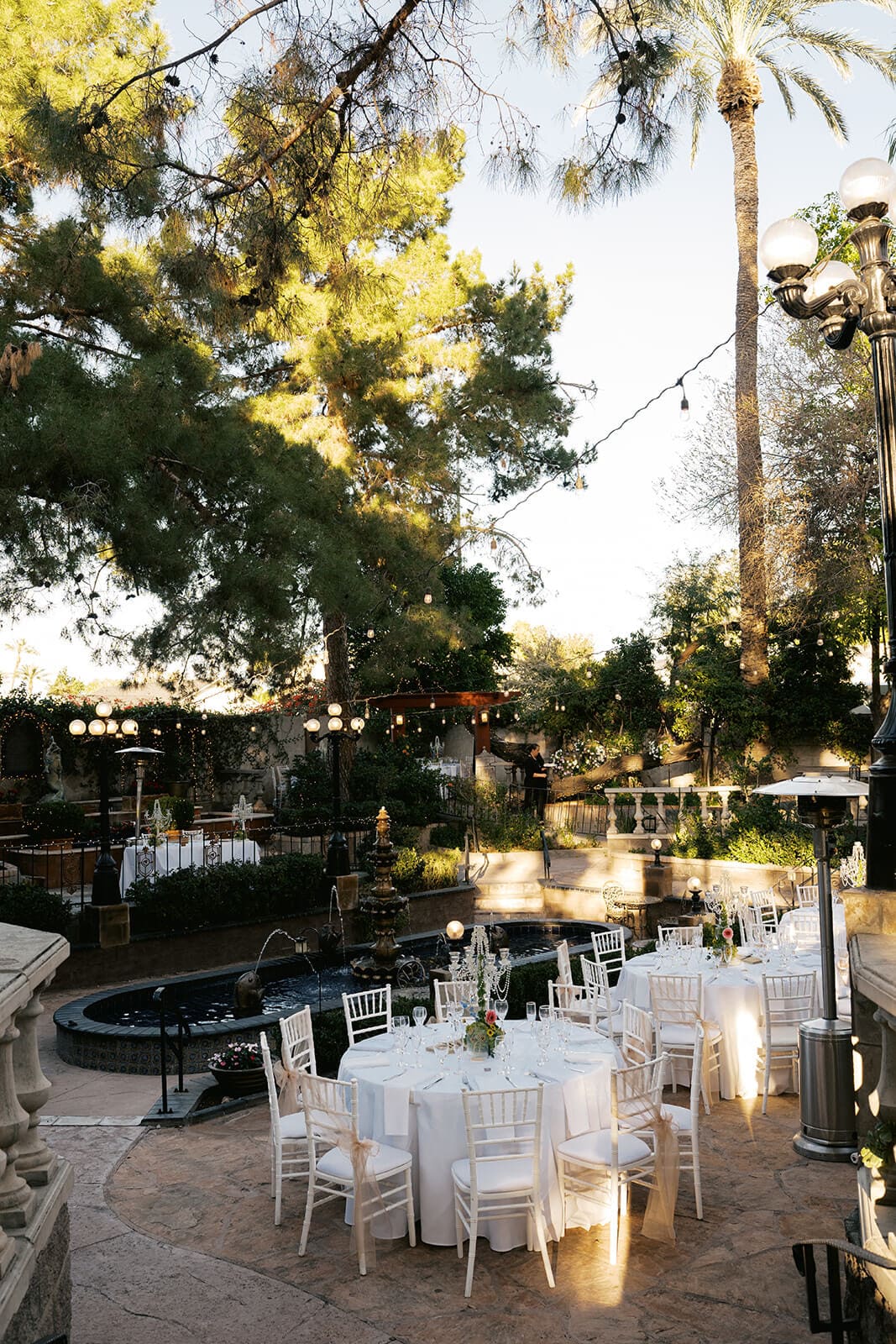 Outdoor reception space at dusk with round tables, chiavari chairs, and string lights among mature trees at Stonebridge Manor garden wedding venue in Mesa Arizona