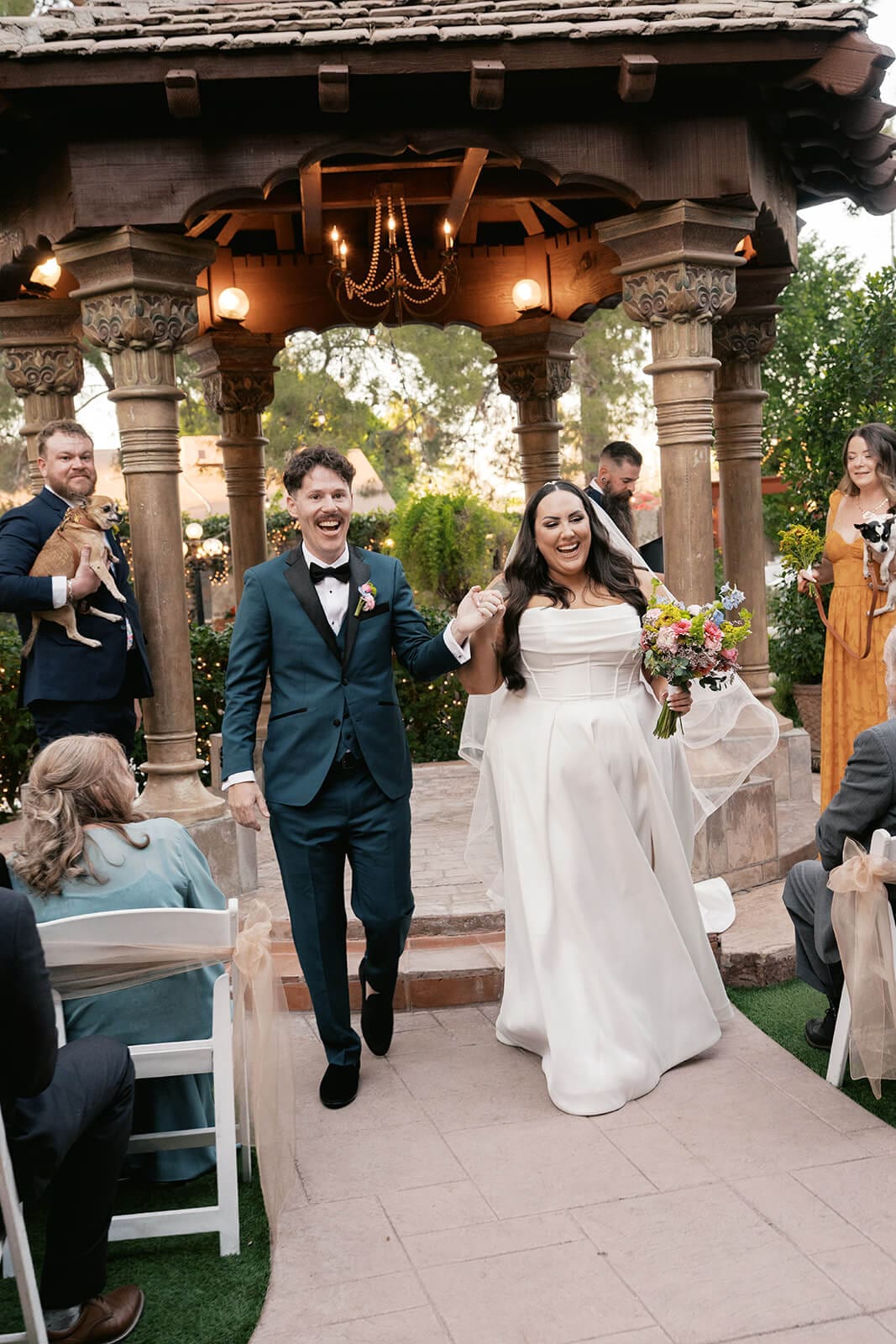 Bride and groom walking back down the aisle laughing under the ornate gazebo with guests cheering at Stonebridge Manor garden wedding venue in Mesa Arizona