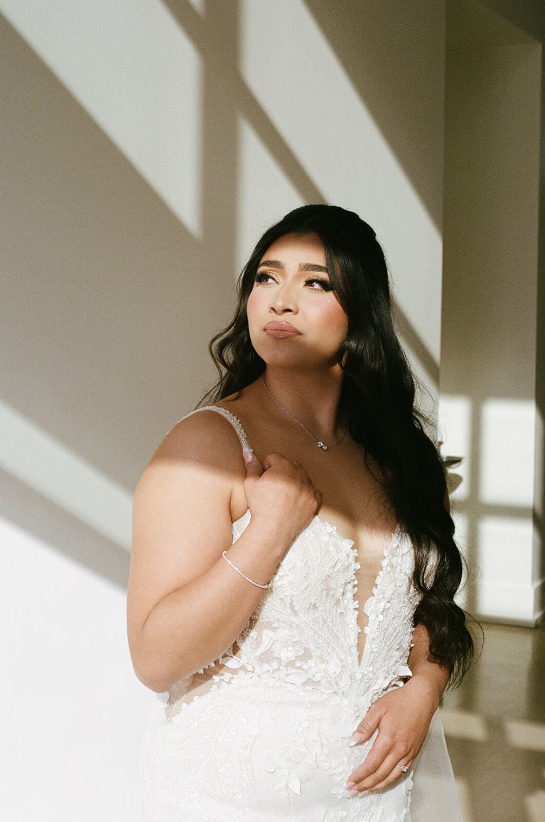 Bride posing in her lace wedding dress as geometric window shadows fall across the wall.