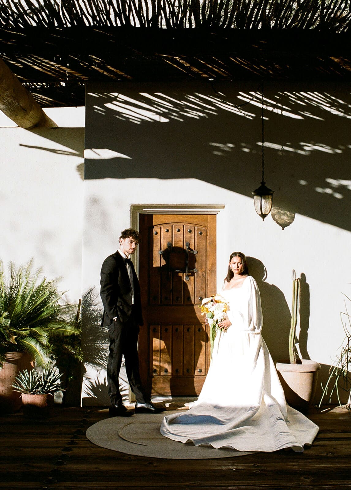 Bride and groom standing in dramatic sunlight outside a Southwestern Airbnb wedding venue with a wooden door and desert plants.
