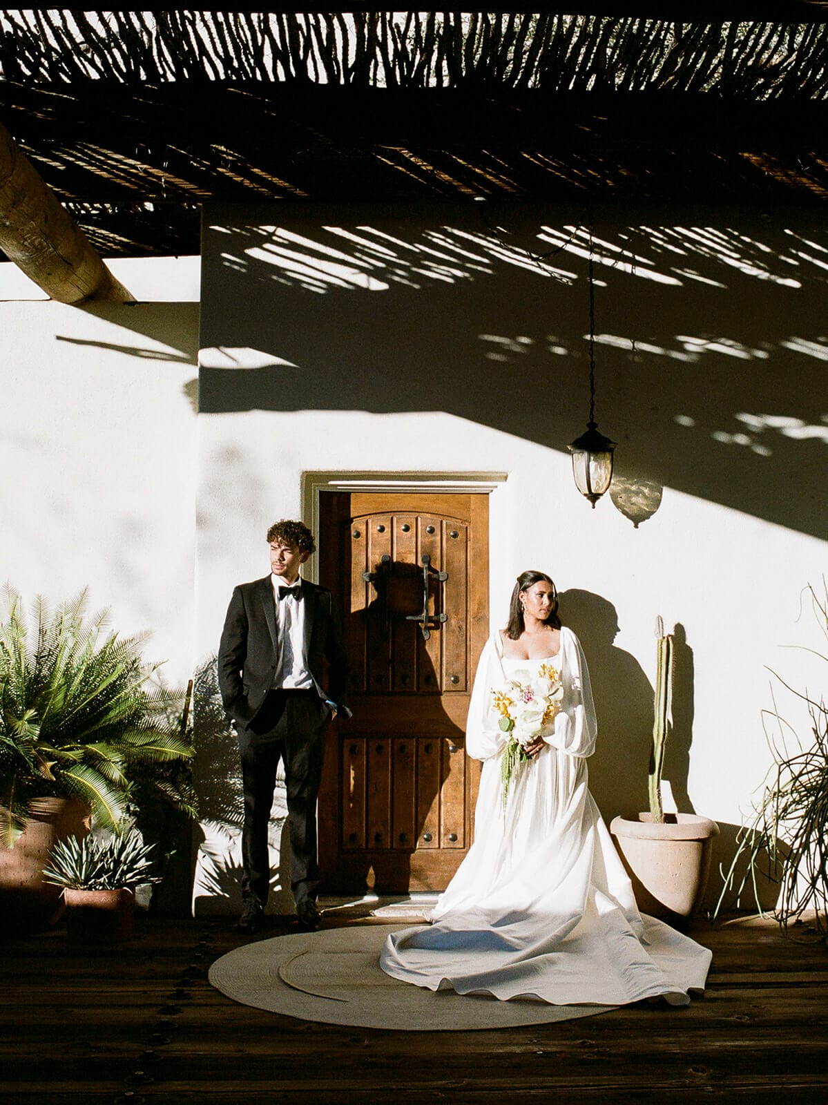 Bride and groom standing in dramatic light outside a desert Airbnb wedding venue with a wooden door and textured shadows.