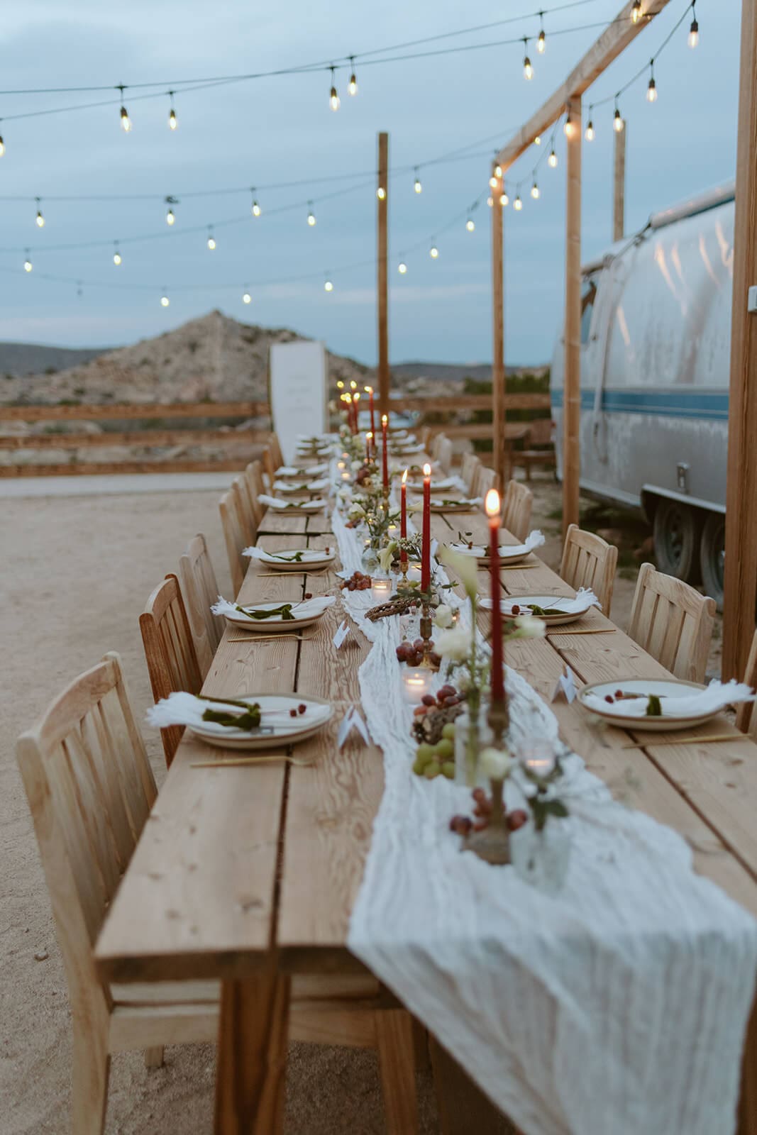 Outdoor desert wedding reception table set with candles, greenery, and string lights beside a vintage trailer.