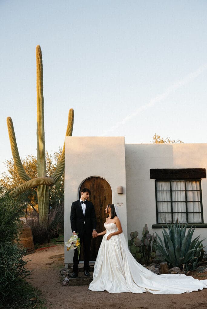 Bride and groom posing outside Joshua Tree House with a tall saguaro cactus and Southwestern architecture behind them.