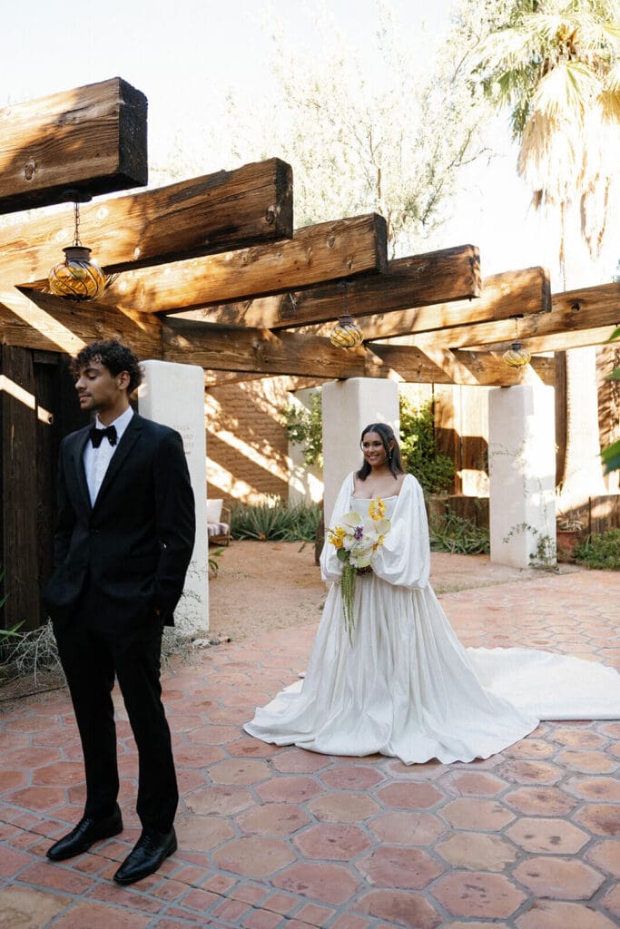Bride and groom standing beneath wooden beams in the courtyard at Joshua Tree House for their desert Airbnb wedding portraits.