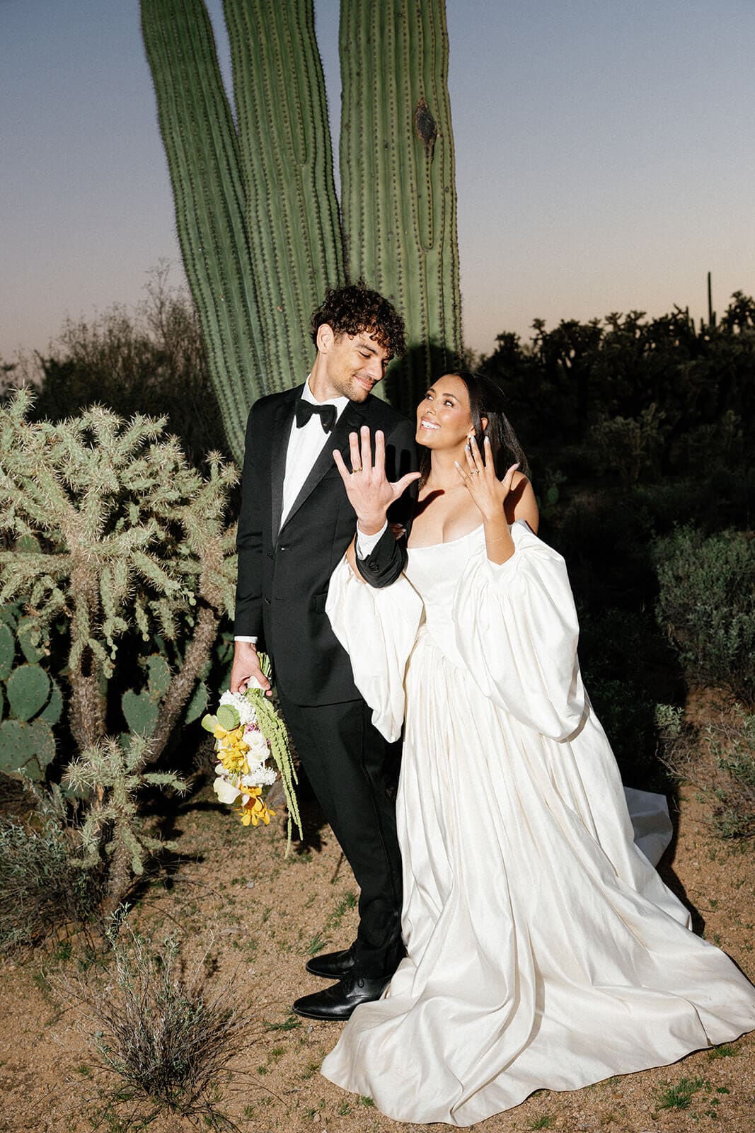 Bride and groom showing off their wedding rings while posing in front of a tall saguaro cactus.