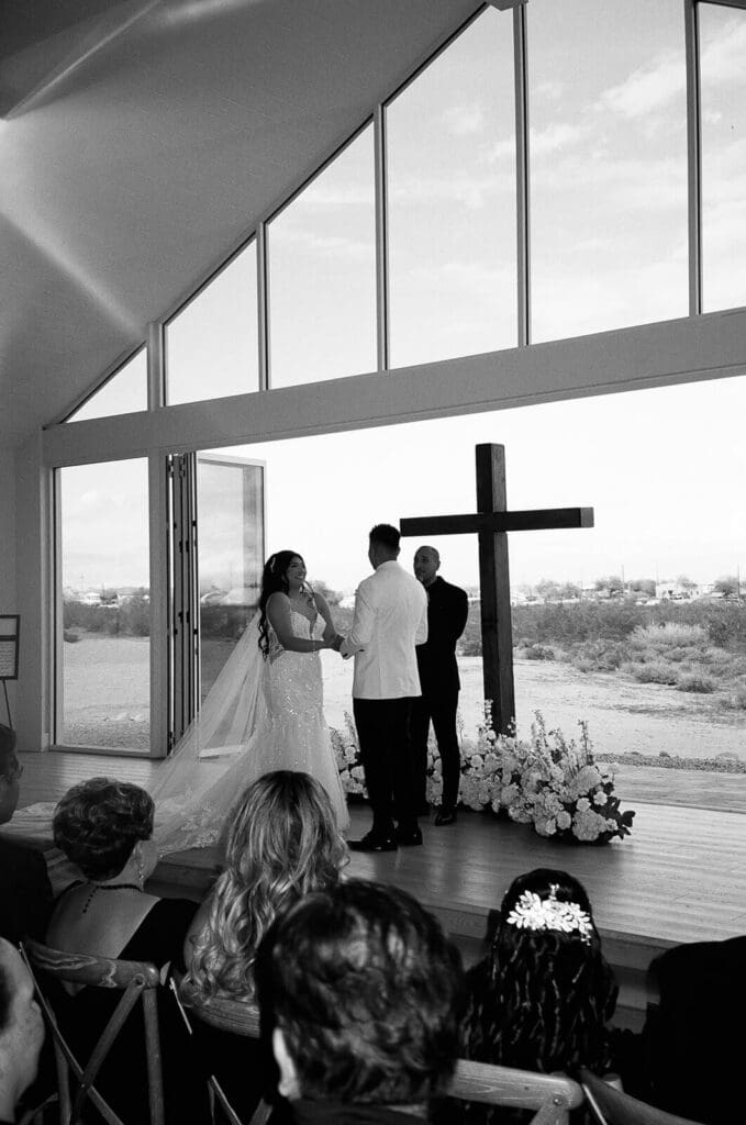 Black-and-white wide view of a wedding ceremony inside a modern glass chapel with a large cross at the altar.