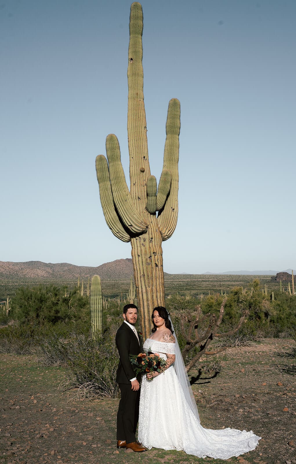 Bride and groom standing beneath a towering saguaro cactus in the Arizona desert.