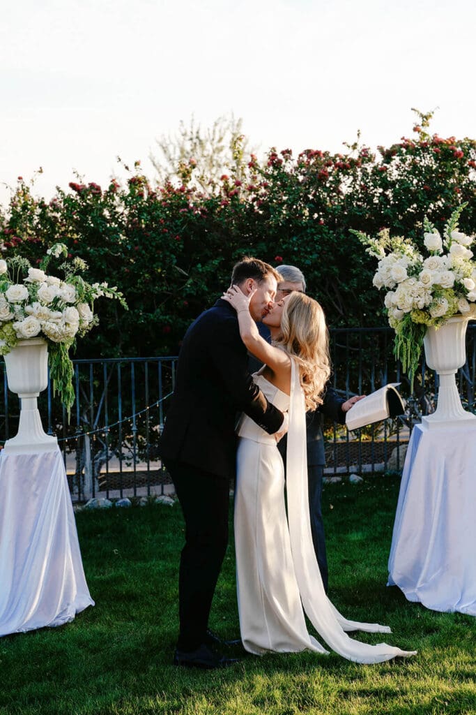 Bride and groom kissing between white floral arrangements during their outdoor Airbnb wedding ceremony.