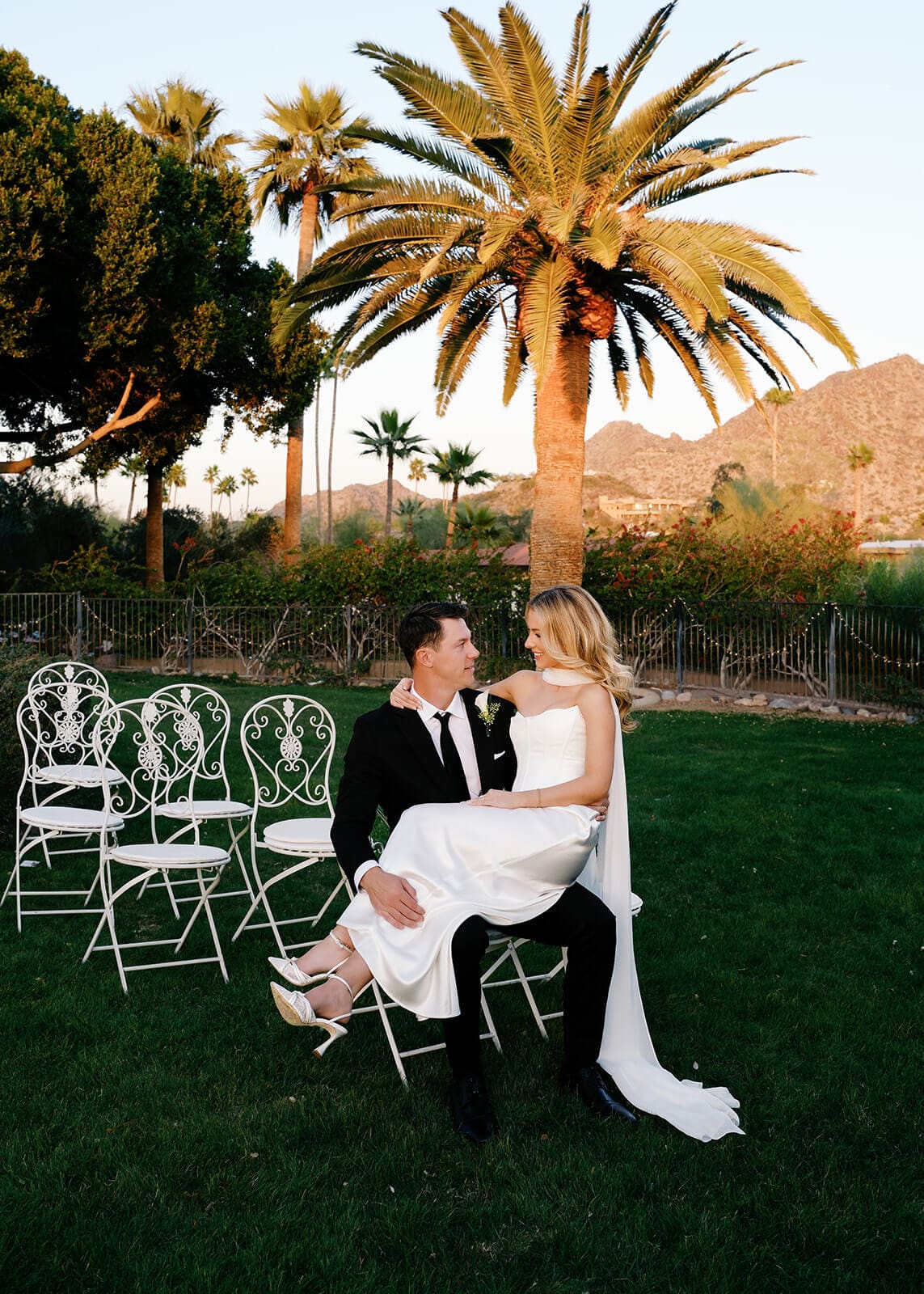 Bride sitting on the groom’s lap beneath palm trees at an outdoor Arizona wedding venue.